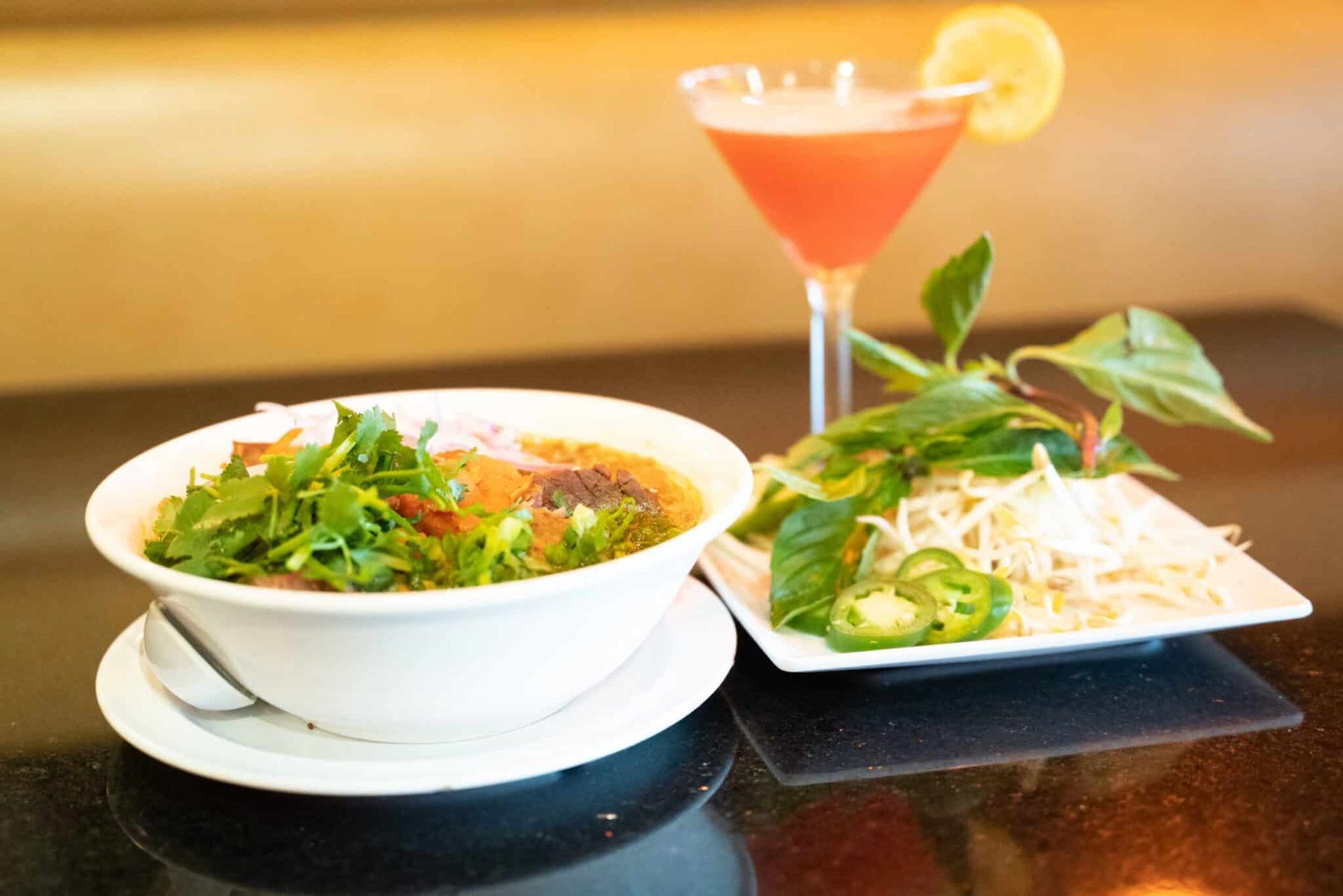 Thai dishes and a pink cocktail on a black table, with herbs and lime garnish