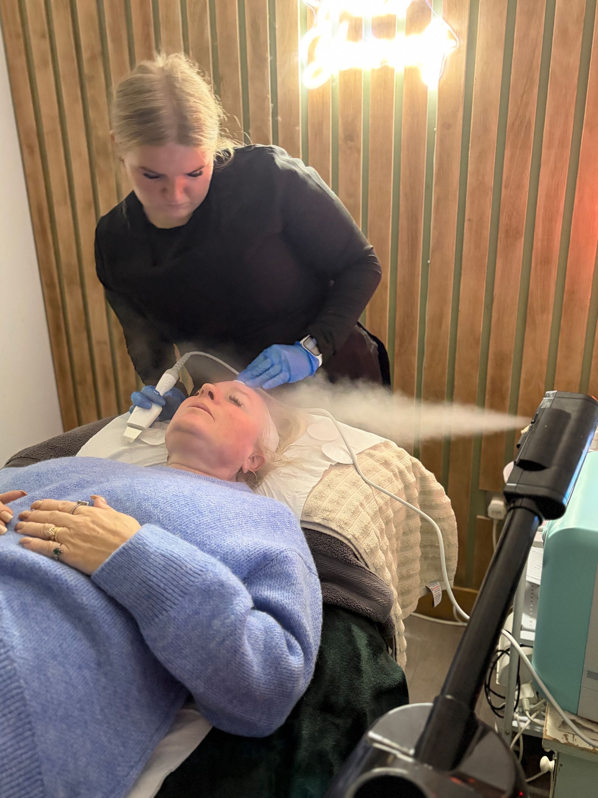 Therapist treating a client’s face with a handheld facial device while a steamer directs steam at her skin