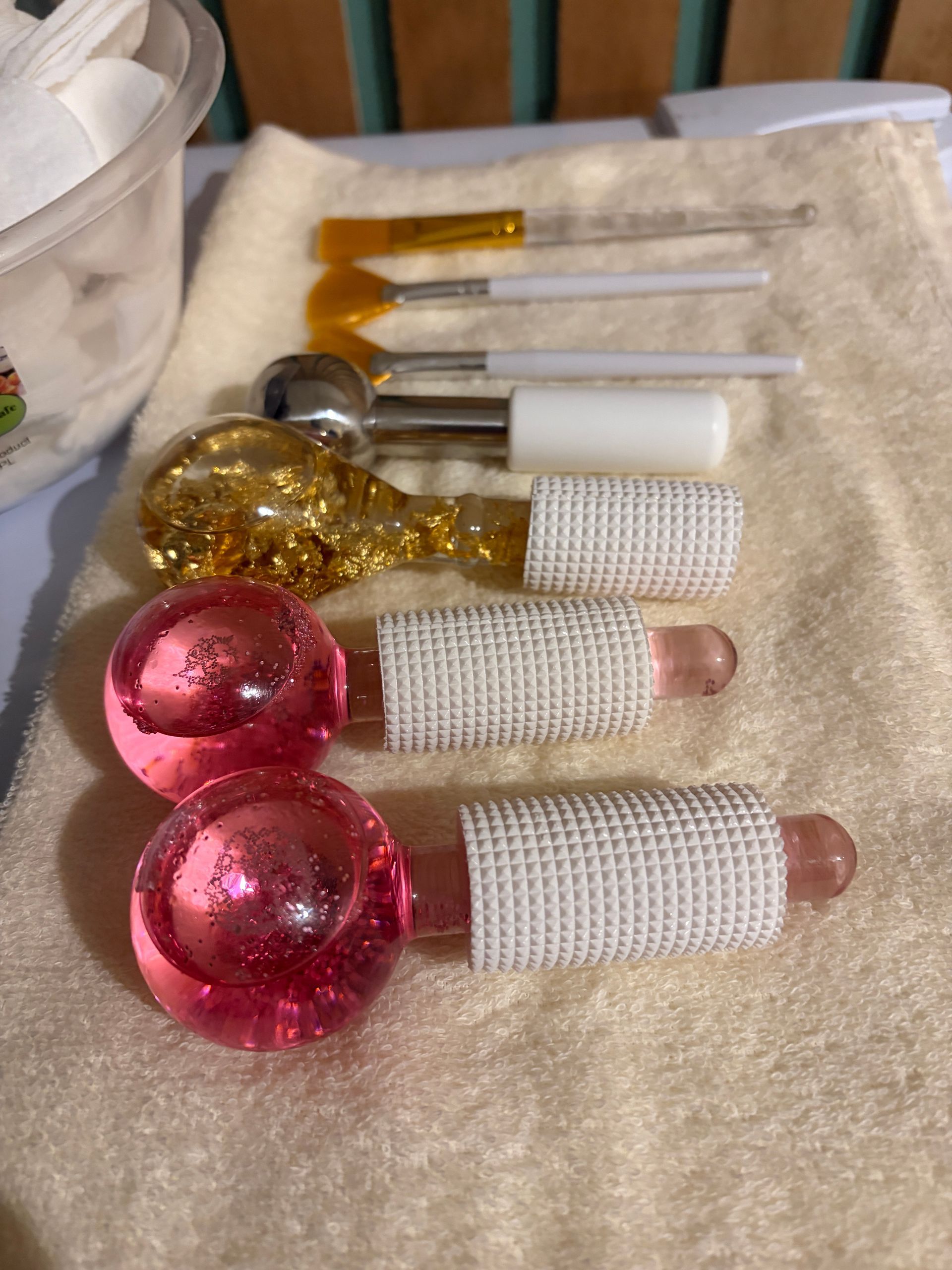 Facial tools laid on a towel, including pink and gold cooling globes, brushes and a metal spoon applicator