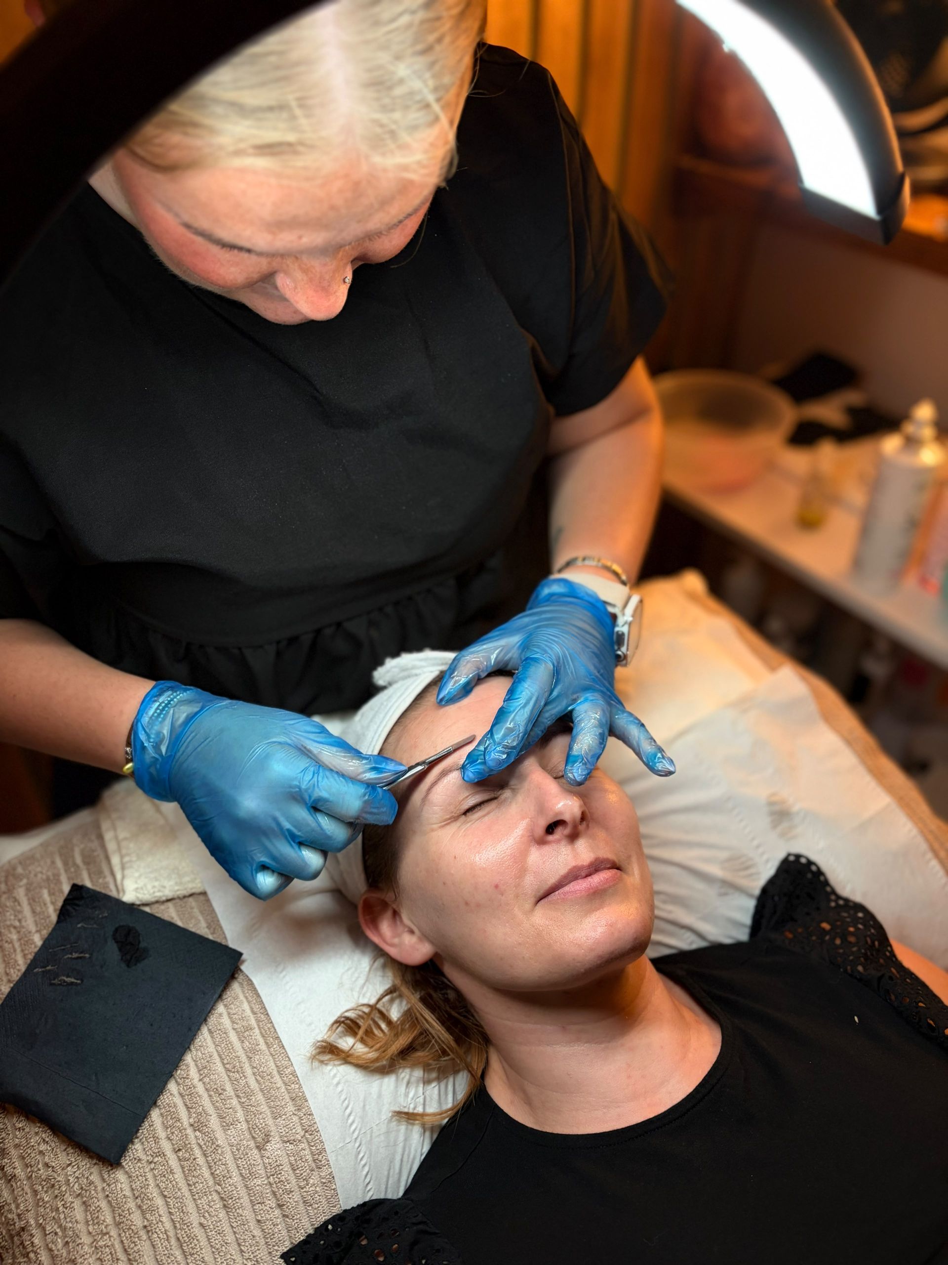 Therapist in blue gloves dermaplaning a client’s forehead with a black card beside her showing the hair removed