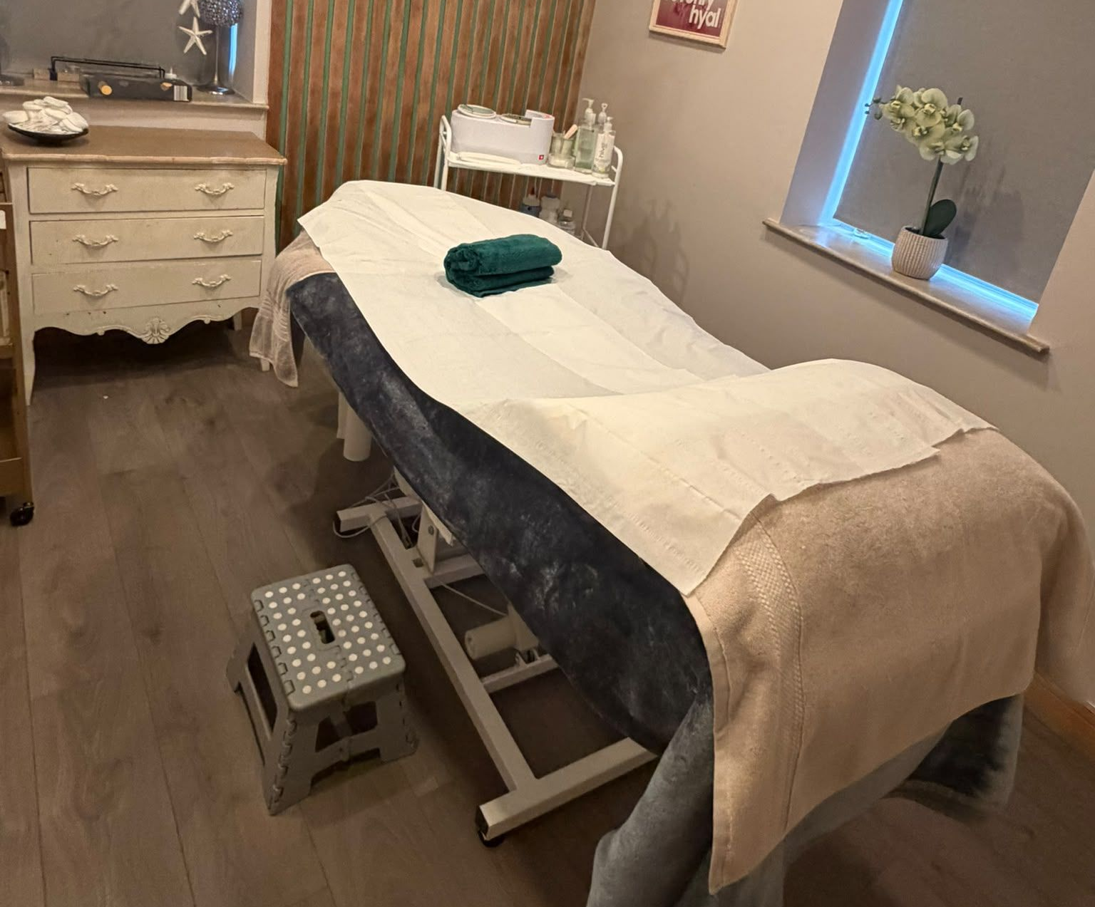Treatment room at Claws Nail and Beauty with a made-up treatment bed, green towel, wood slat feature wall and a waxing trolley