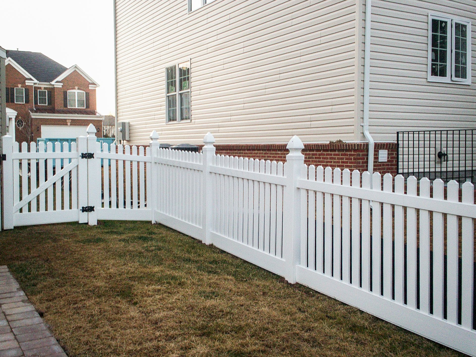 White picket fence bordering a yard with brown grass, next to a beige house.