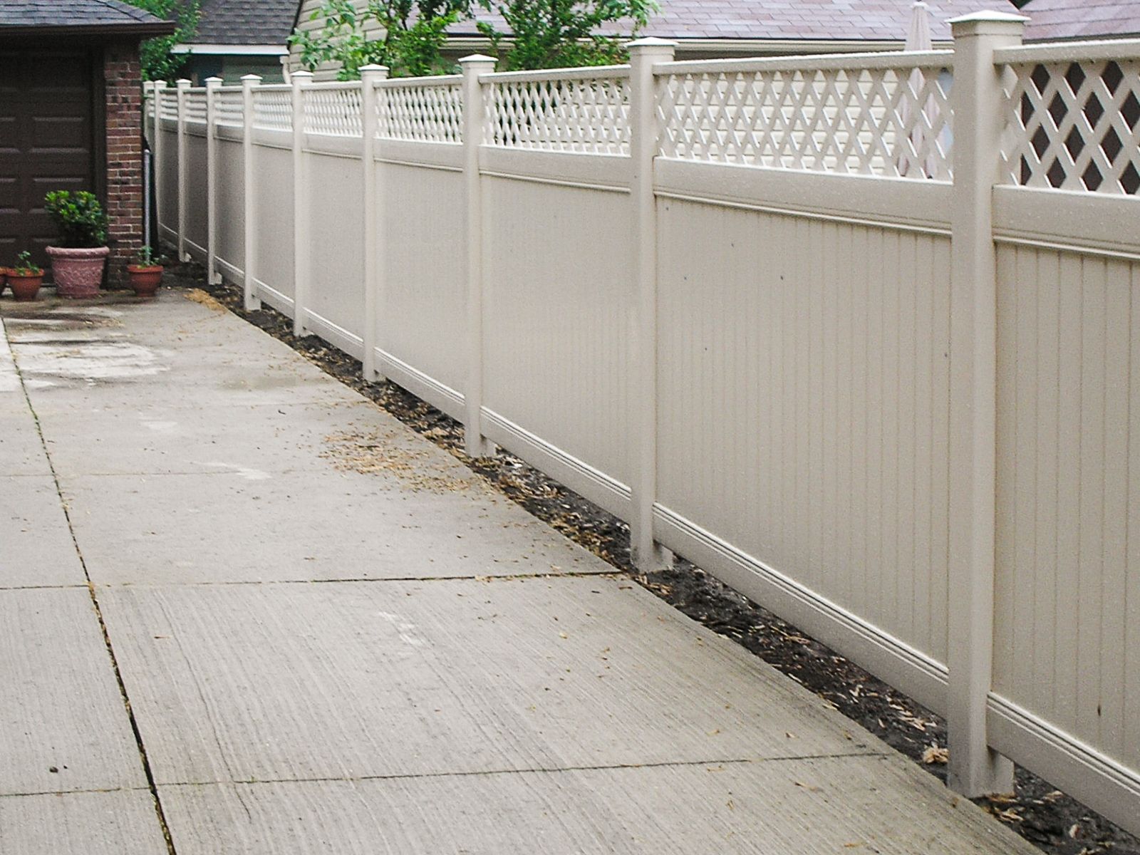 Beige vinyl privacy fence along a concrete driveway, topped with lattice.