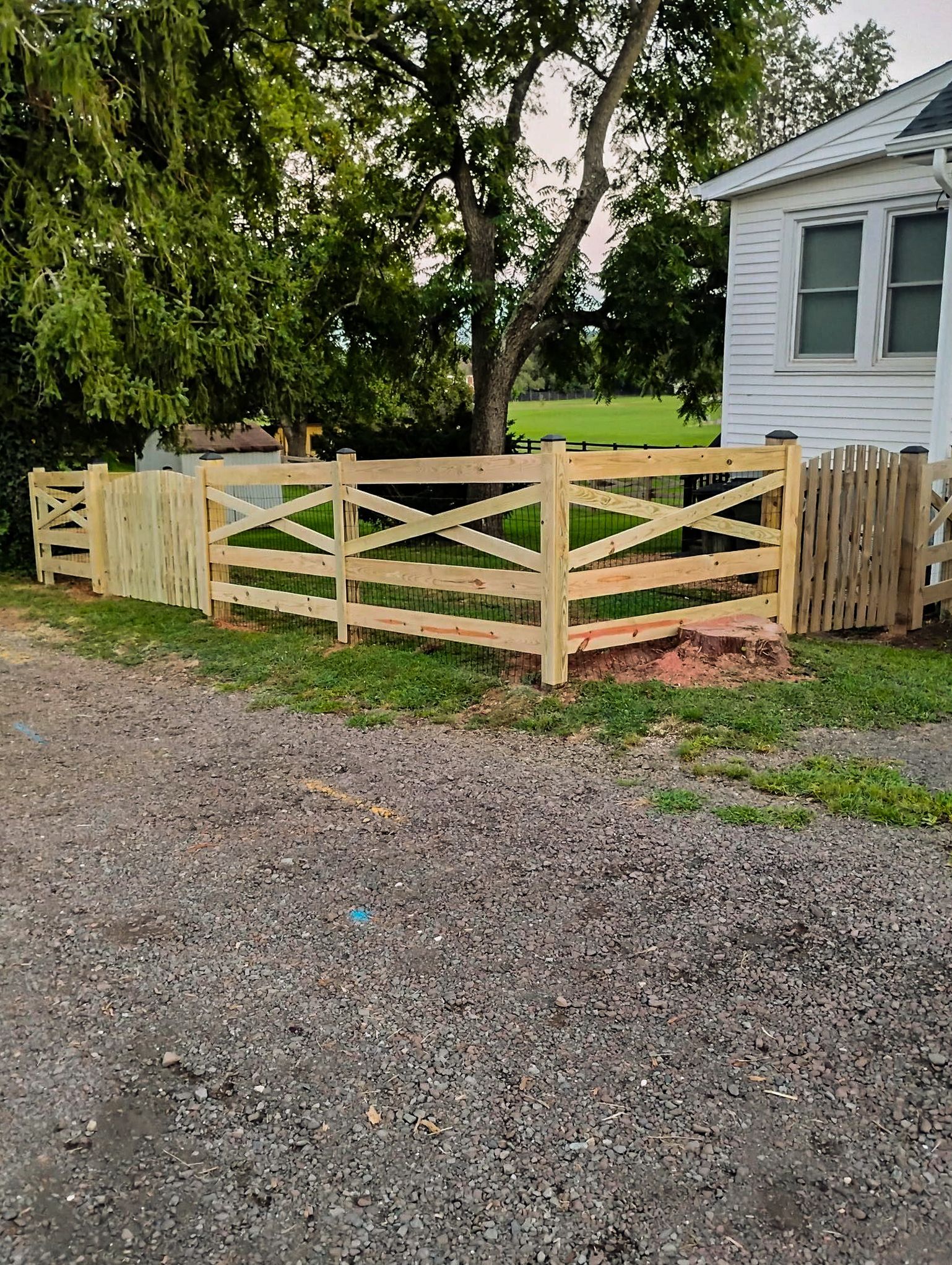 Wooden fence with an X design, in front of a white building with windows. Gravel ground.