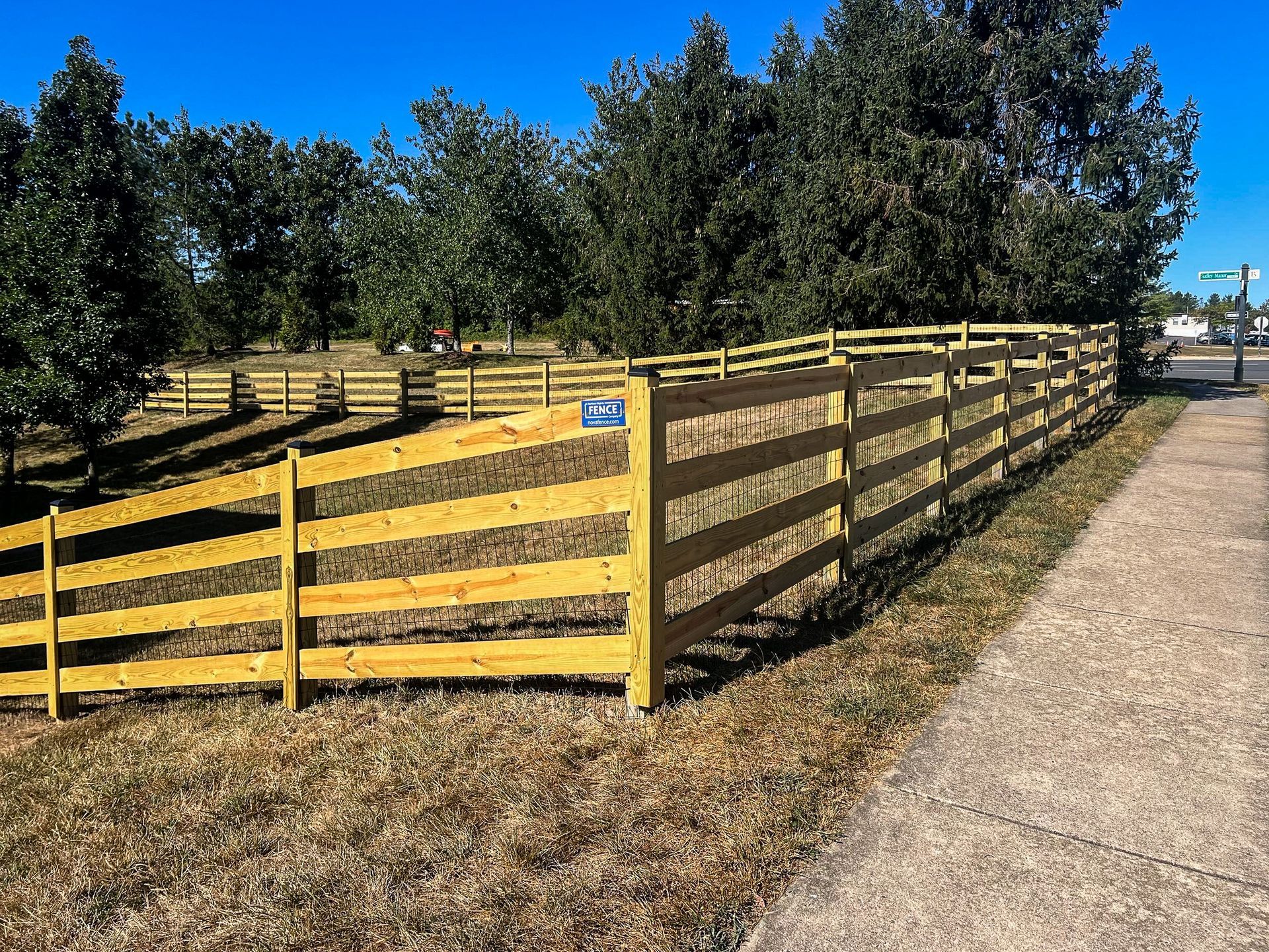 Wooden fence bordering a grassy area next to a sidewalk and road, under a blue sky.