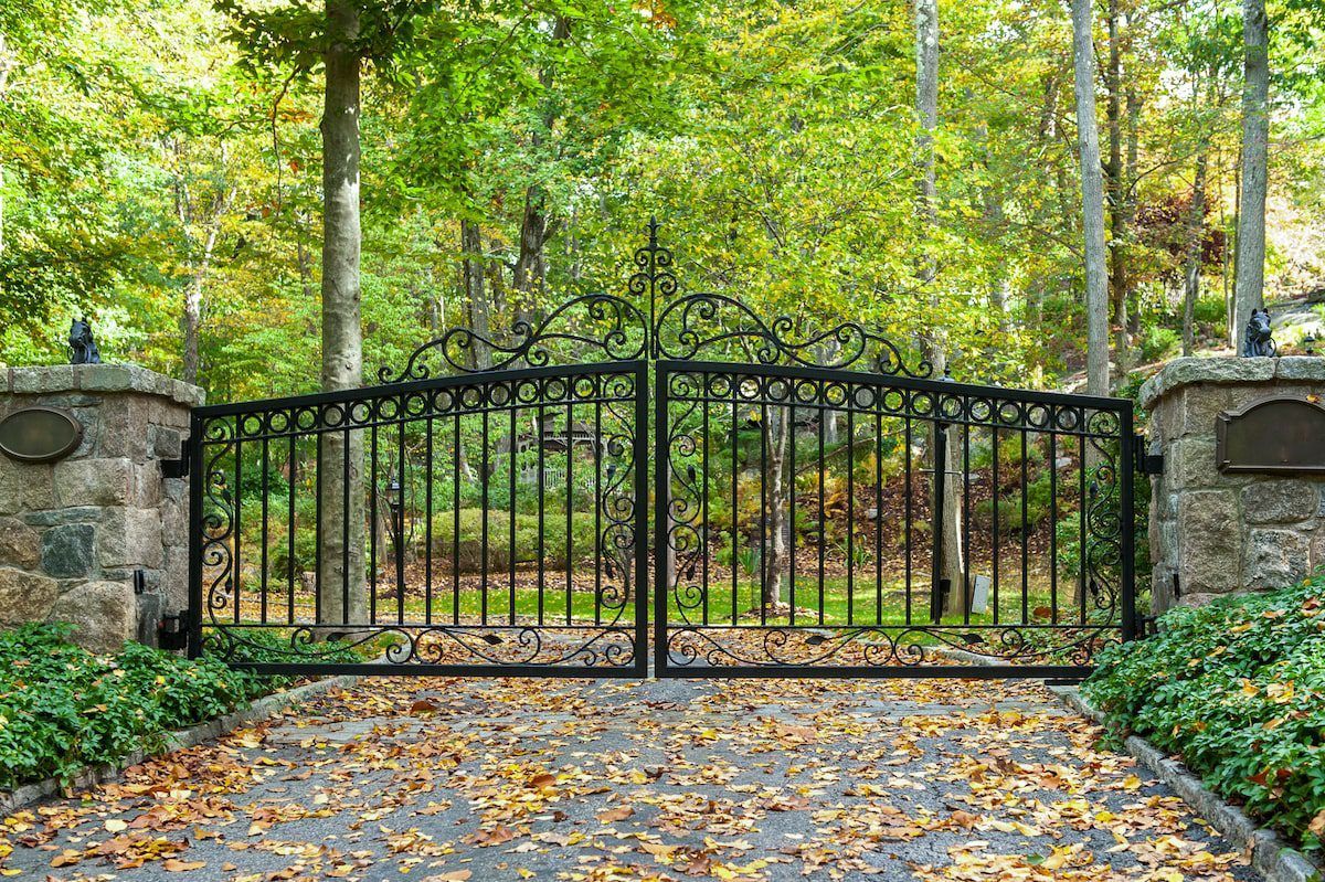 Black wrought iron gate set between stone pillars, leading into a wooded area. Autumn leaves on the driveway.