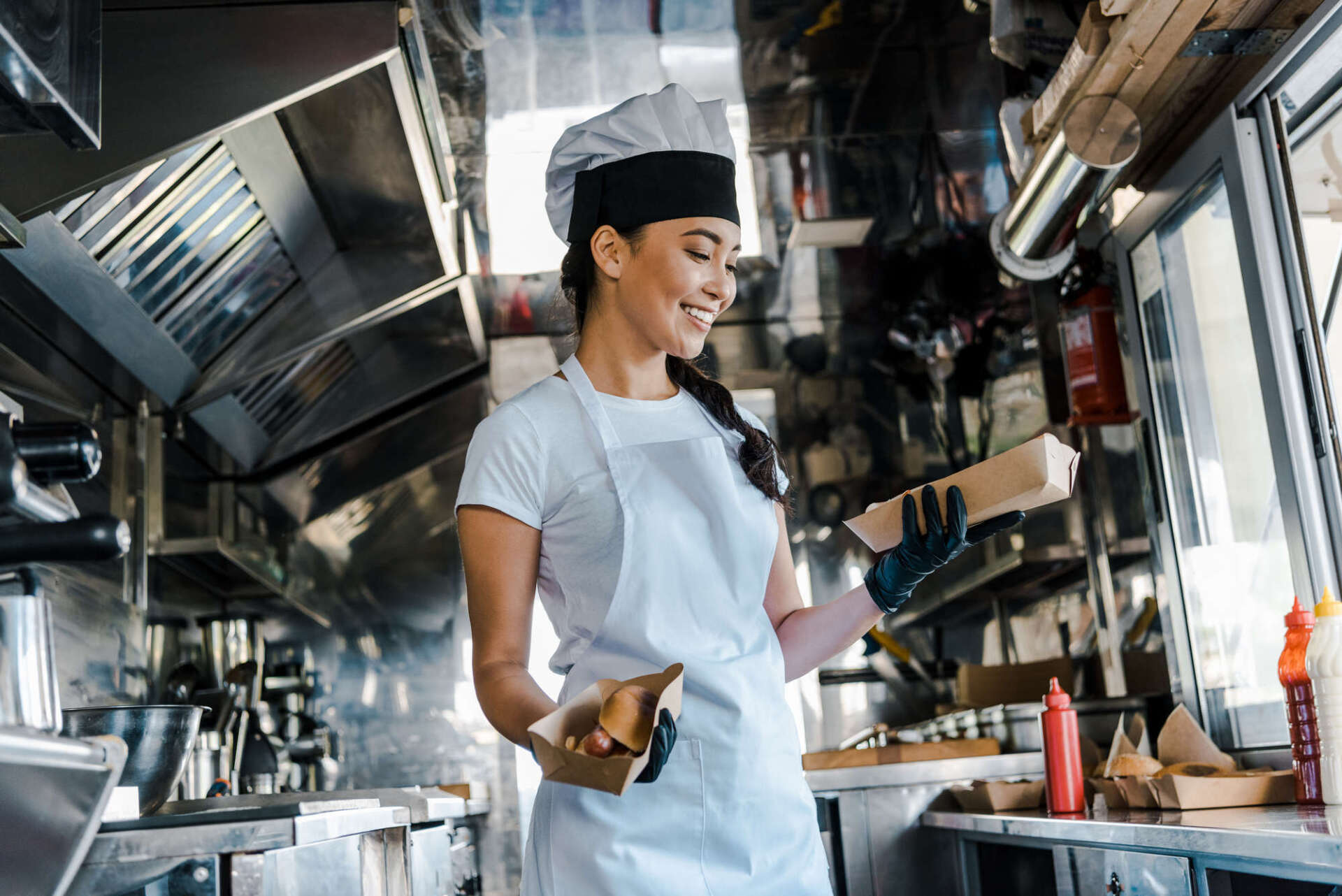 Woman in chef hat and apron in food truck, holding food, smiling.