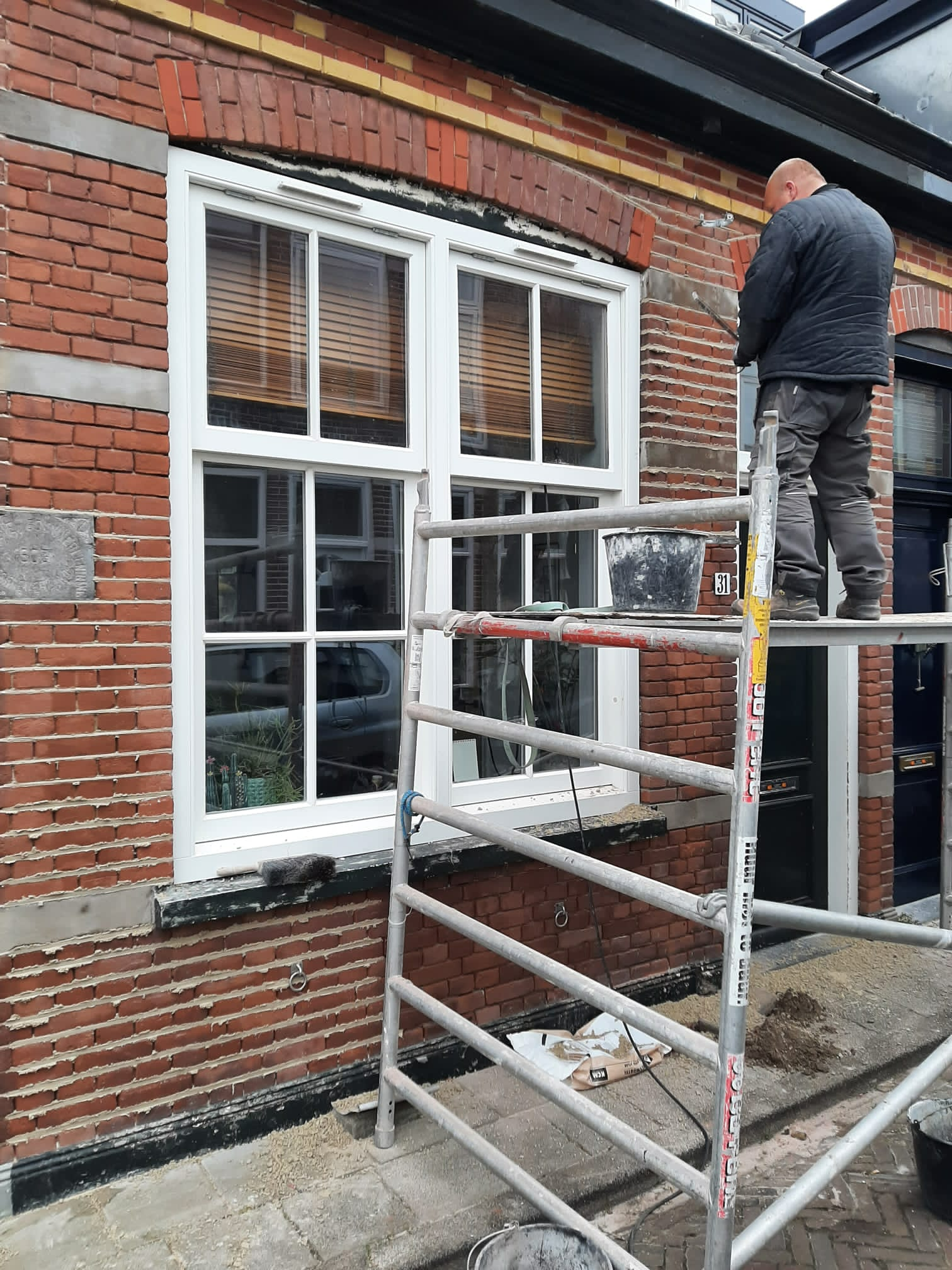 Een arbeider op een steiger installeert een raam met een wit kozijn in een rode bakstenen muur.