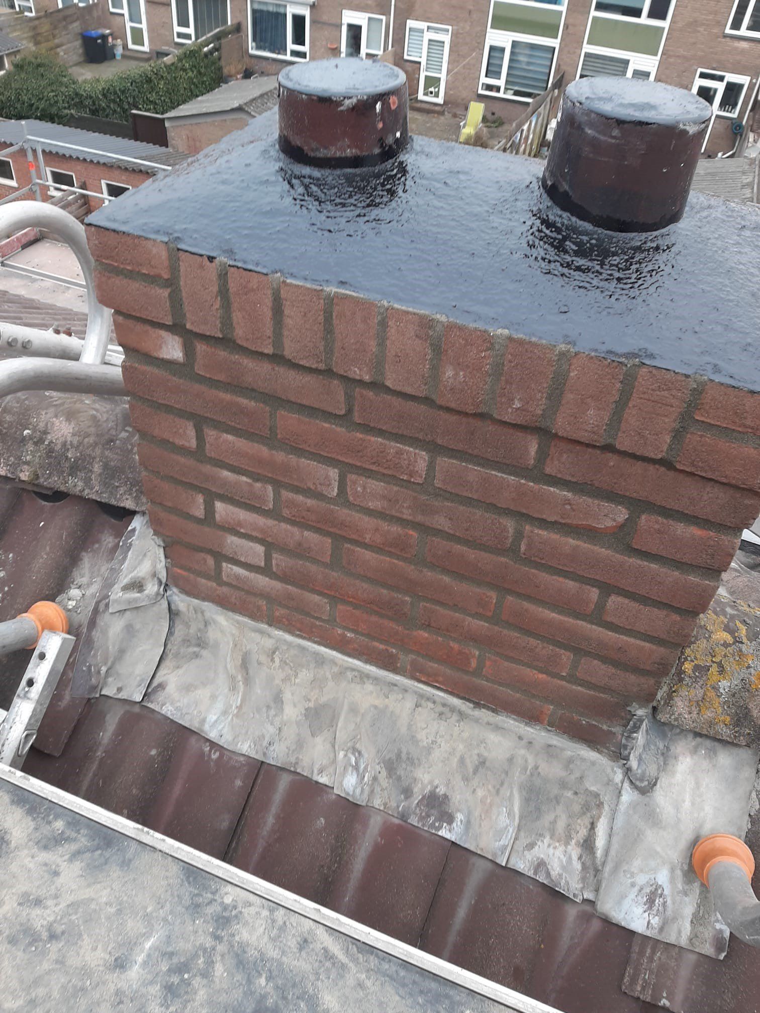 Een bakstenen schoorsteen op een dak met twee zwarte ventilatiekappen en een natte teerafdichting.