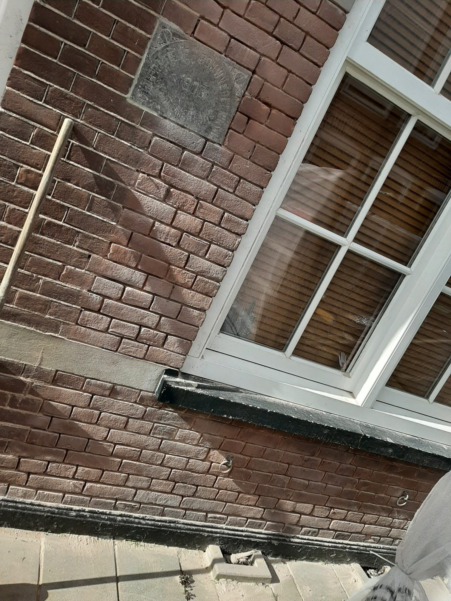 Schuin zicht op een bakstenen gebouwmuur met een raam met witte ruiten en een stenen richel vlakbij een trottoir.