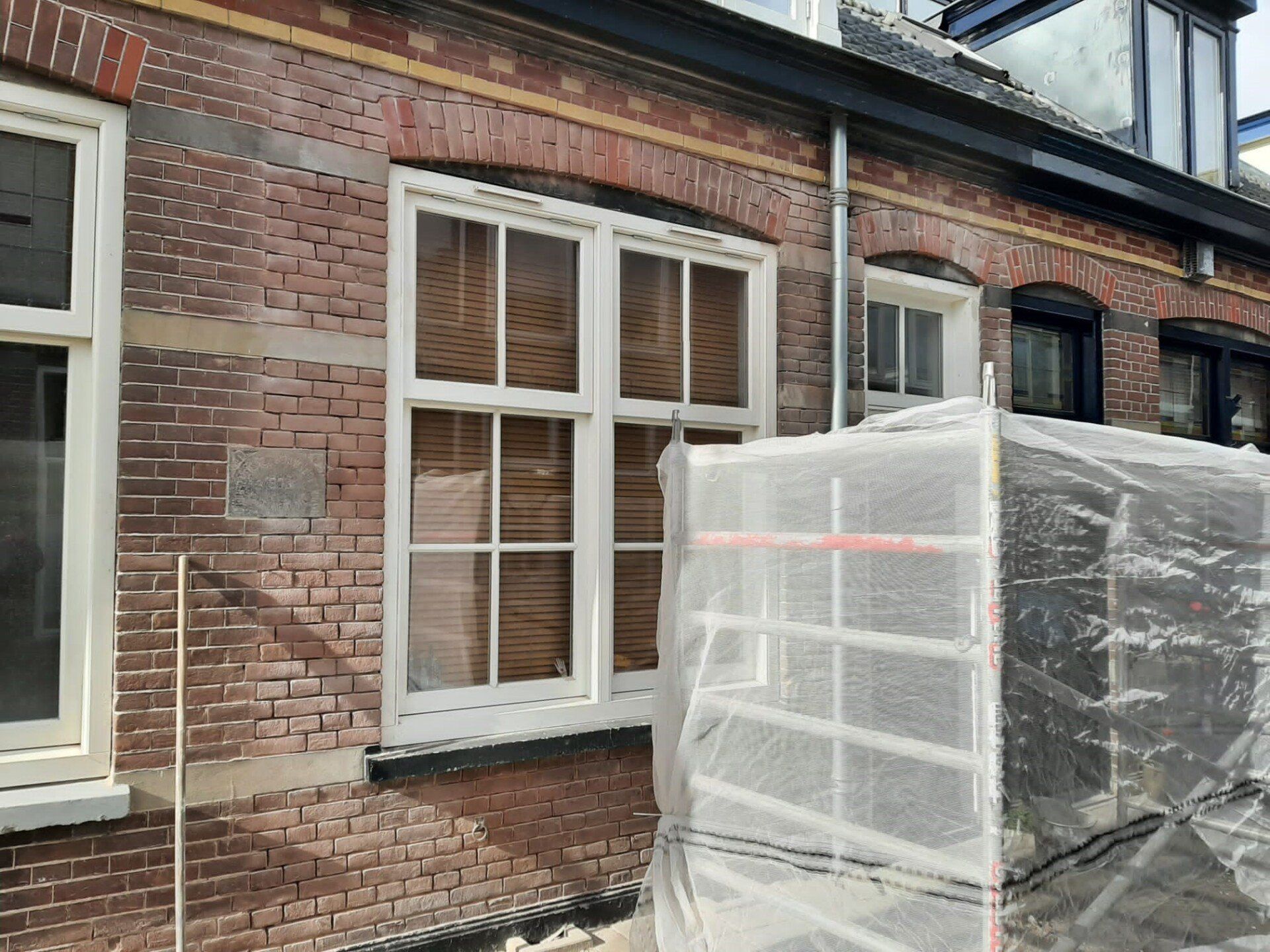 Een bakstenen gevel met witte raamkozijnen en een steiger die met een plastic zeil is omwikkeld.
