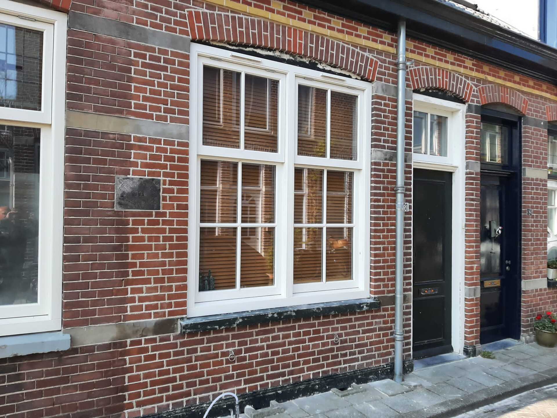 Een bakstenen gevel van een rijtjeshuis met wit omlijste ramen en een donkere, open deuropening langs het trottoir.
