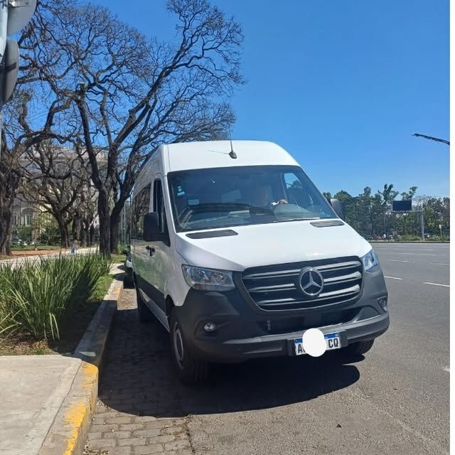 Una furgoneta Mercedes blanca estacionada junto a la acera en un día soleado. Se ven árboles y edificios al fondo.