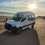 Furgoneta blanca estacionada en una playa de arena con el sol al fondo.