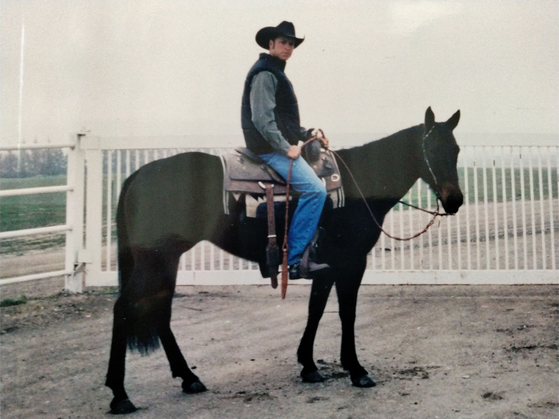 Man on horseback wearing cowboy hat, saddle, and jeans, outside a white fence.