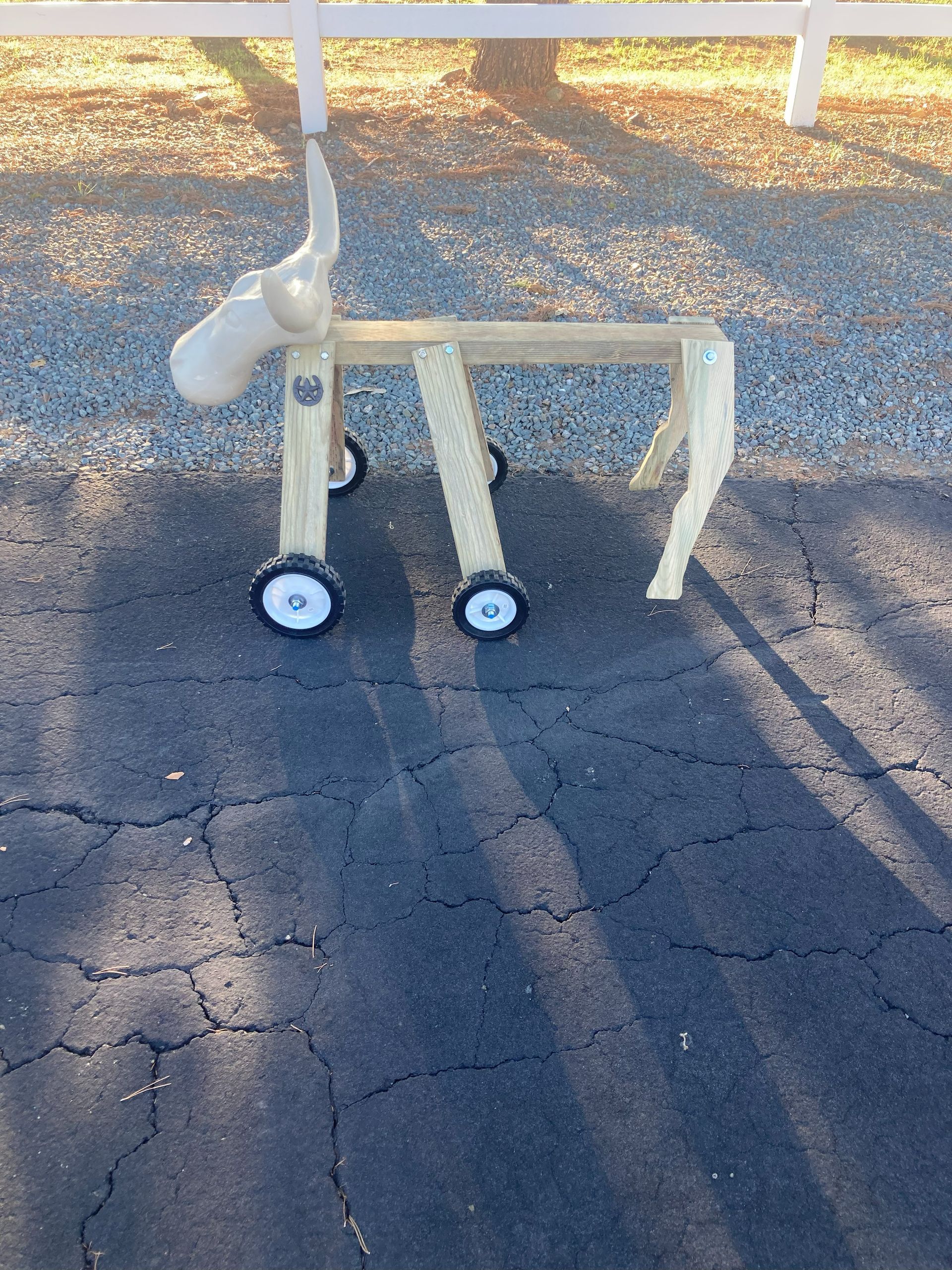 A wooden moose-shaped push toy on wheels standing on an asphalt driveway near a white fence.
