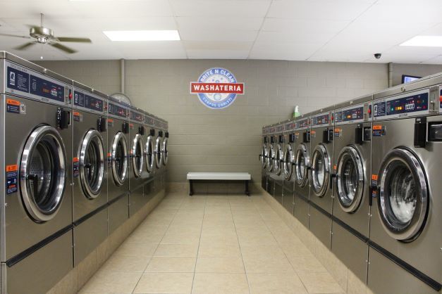 Row of silver front-loading washing machines in a clean laundromat with a bench at the back