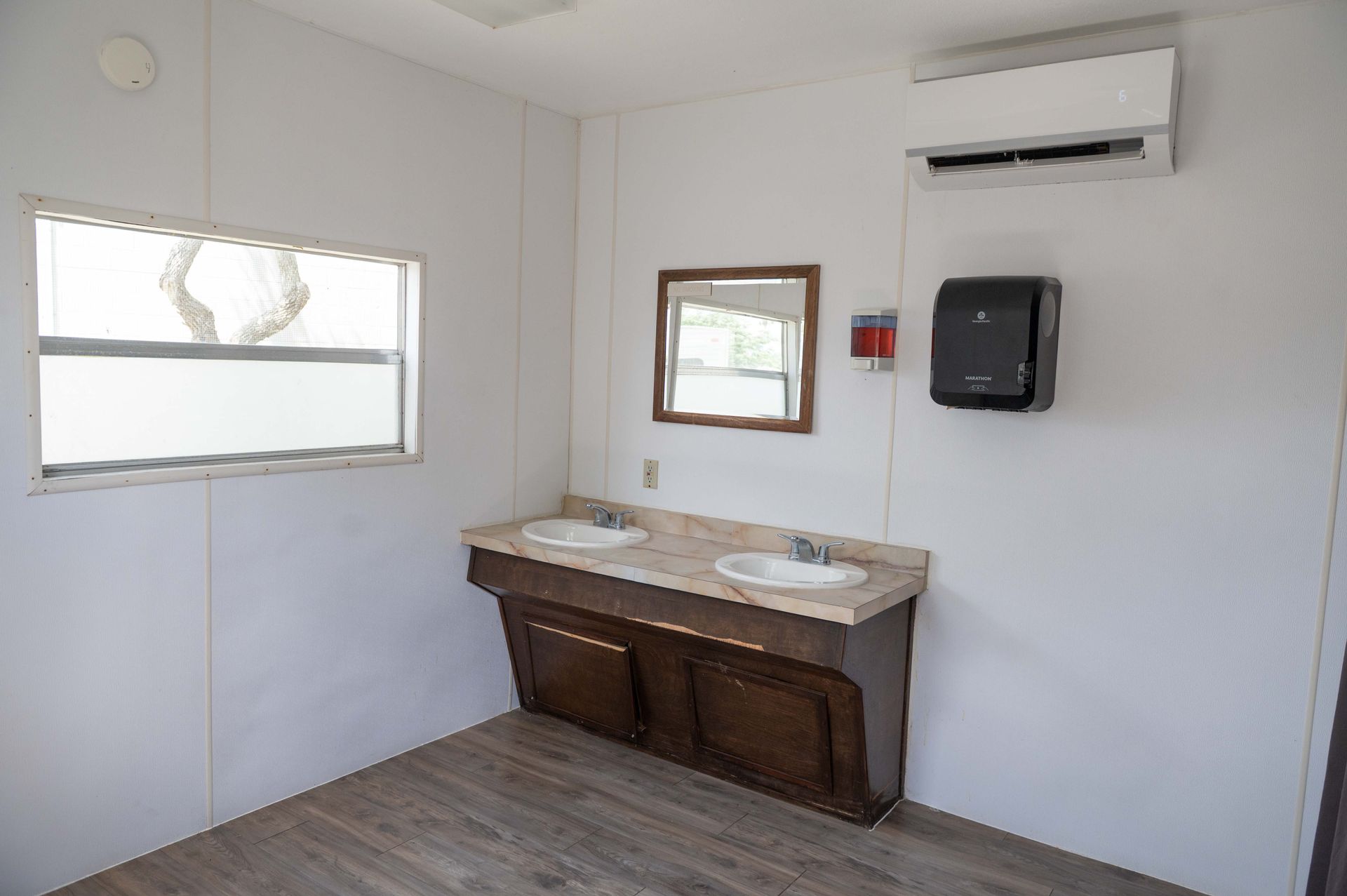 Small public restroom with double sink vanity, mirror, window, and wall-mounted soap dispenser.