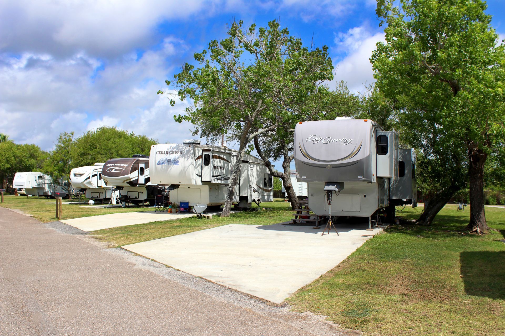 RVs parked at a grassy campground beside a paved site under a blue sky