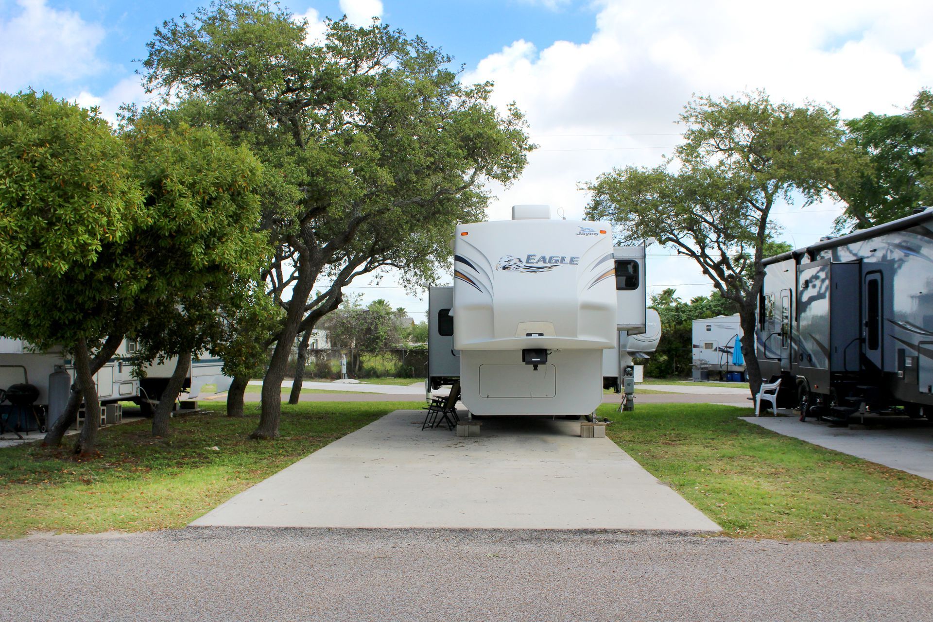 RV parked on a concrete pad among trees and neighboring campers under a partly cloudy sky