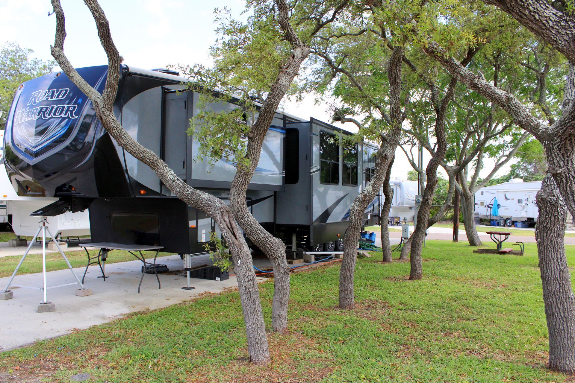 Modern gray travel trailer parked on a grassy campsite among trees, with its awning extended.