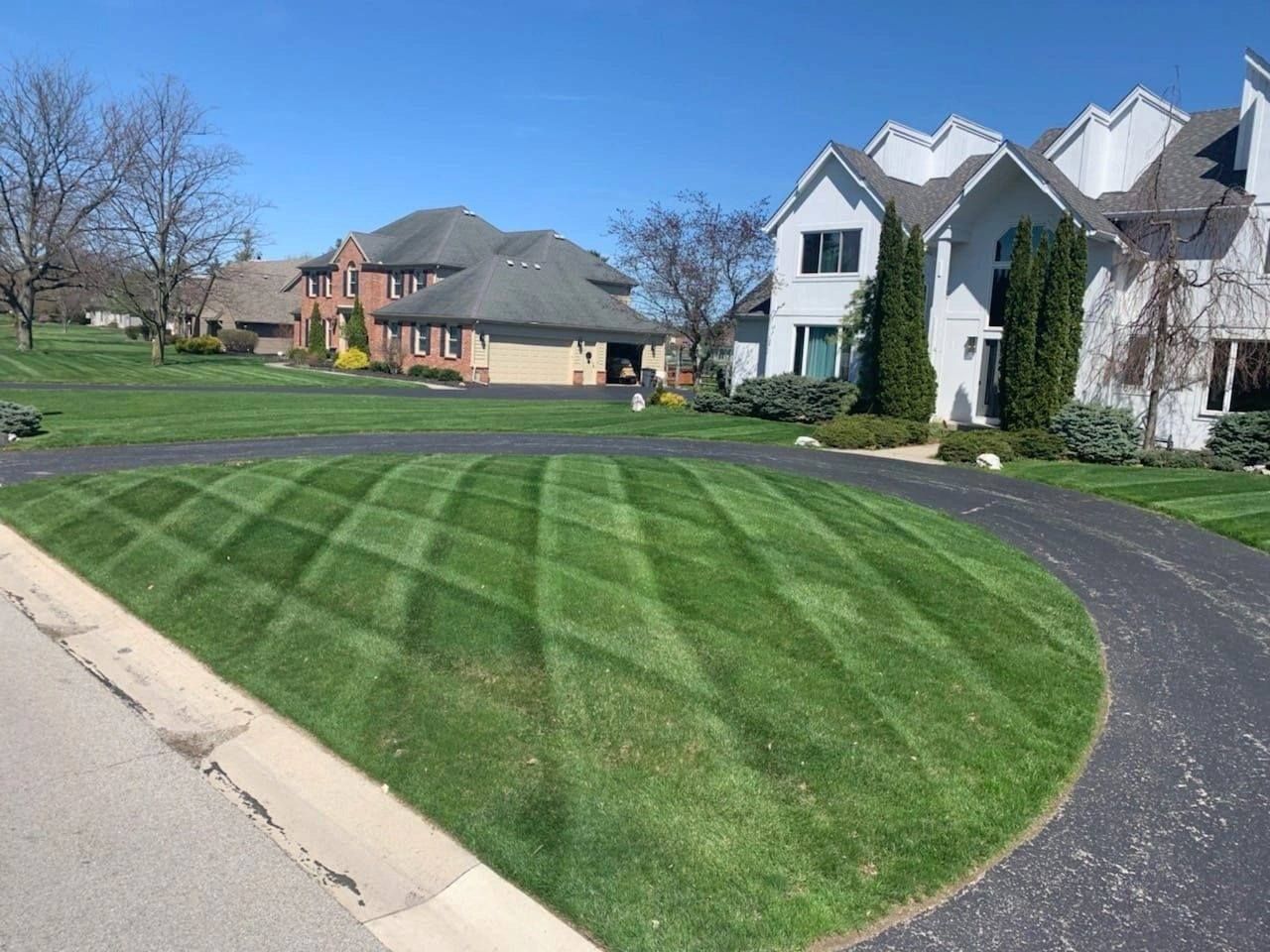 Curved green lawn beside a suburban sidewalk and driveway with houses under a clear blue sky