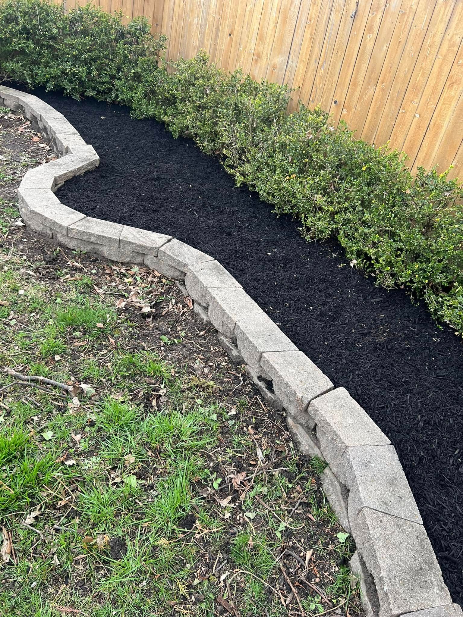 Curved stone garden border separating black mulch and grass beside a wooden fence.