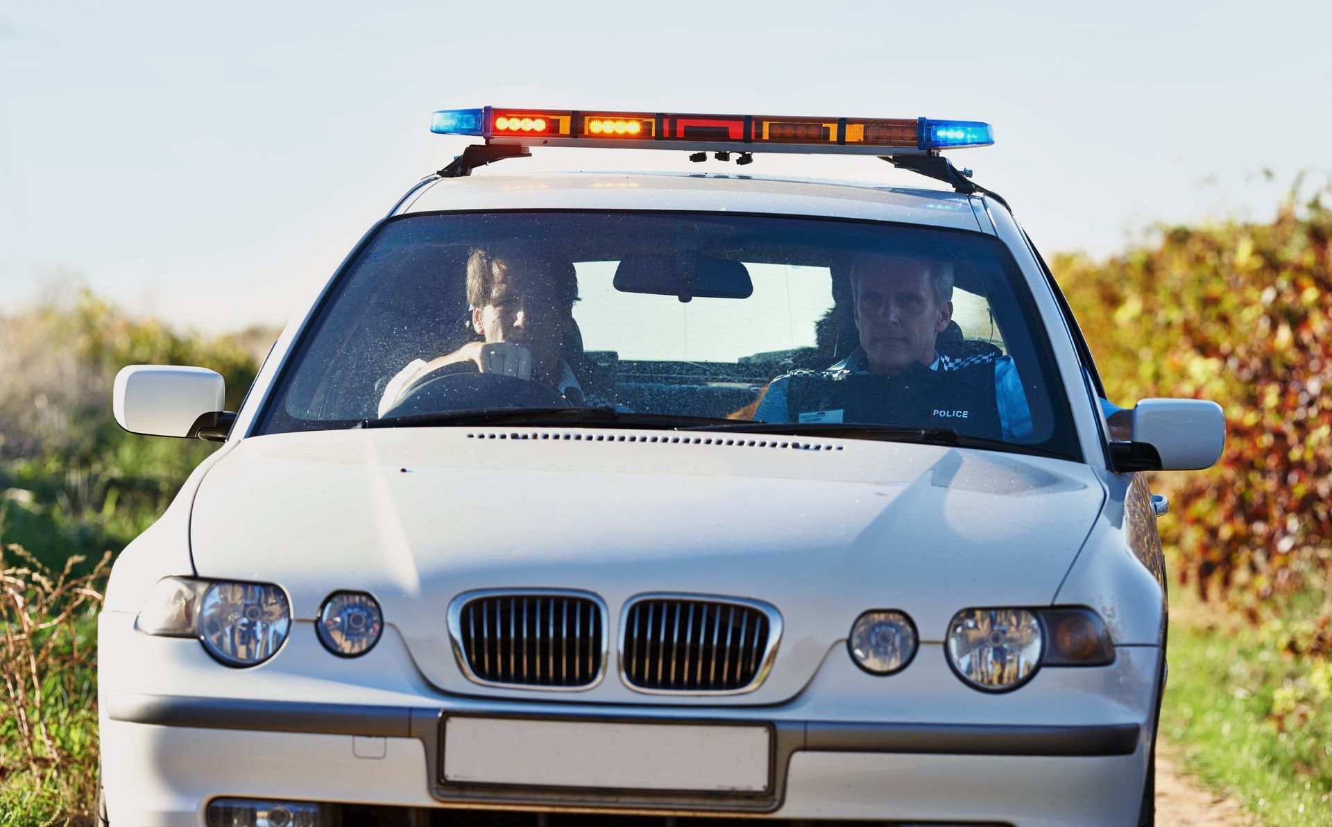 White police car with flashing lights, two officers inside, driving on a road.