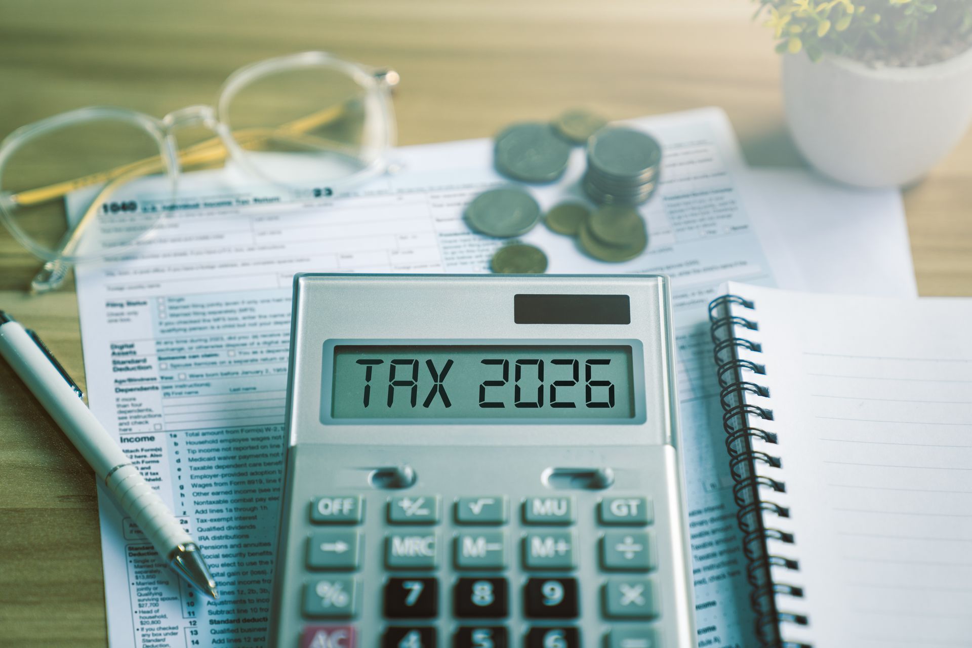 Calculator showing “Tax 2026” with tax forms, coins, pen, and notebook on a desk. Calculator showing “Tax 2026” with tax forms, coins, pen, and notebook on a desk.