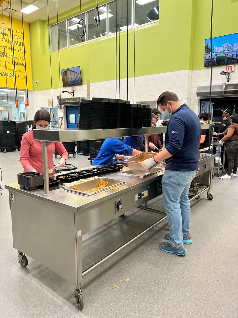 People assembling food in a commercial kitchen, wearing masks, with a long stainless steel counter.