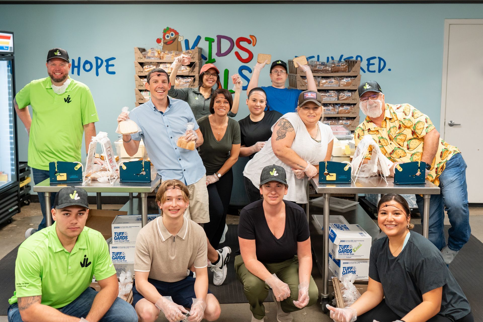 Group of people packing food at a "Kids Meals Delivered" facility. They're smiling and appear to be volunteers.