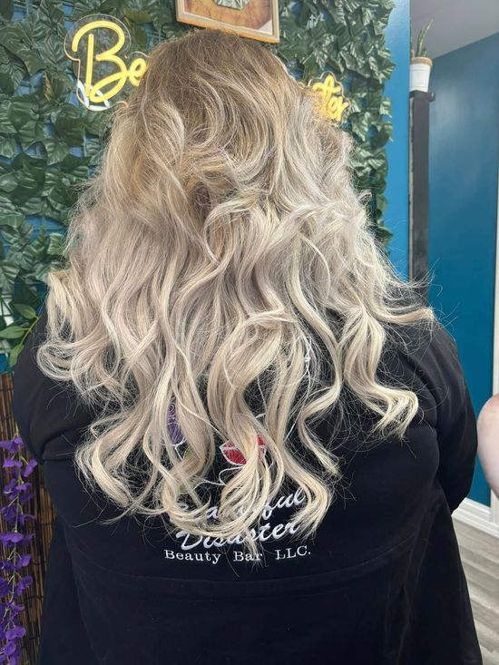 Back view of a person with long wavy blond hair in a salon chair, against a leafy wall and blue accent wall