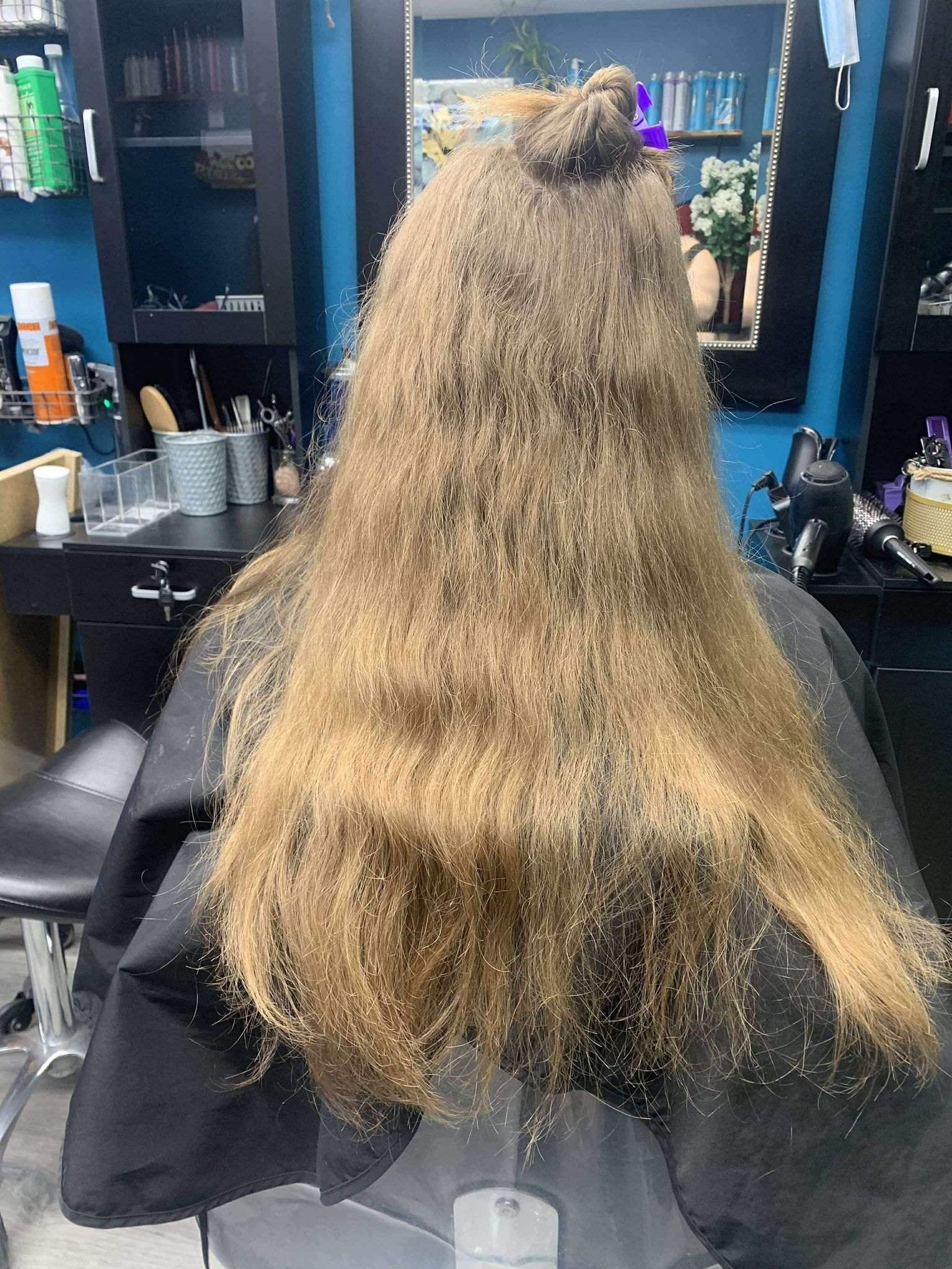 Person with long curly hair sitting in a salon chair, viewed from behind, in front of mirrors and hair products.