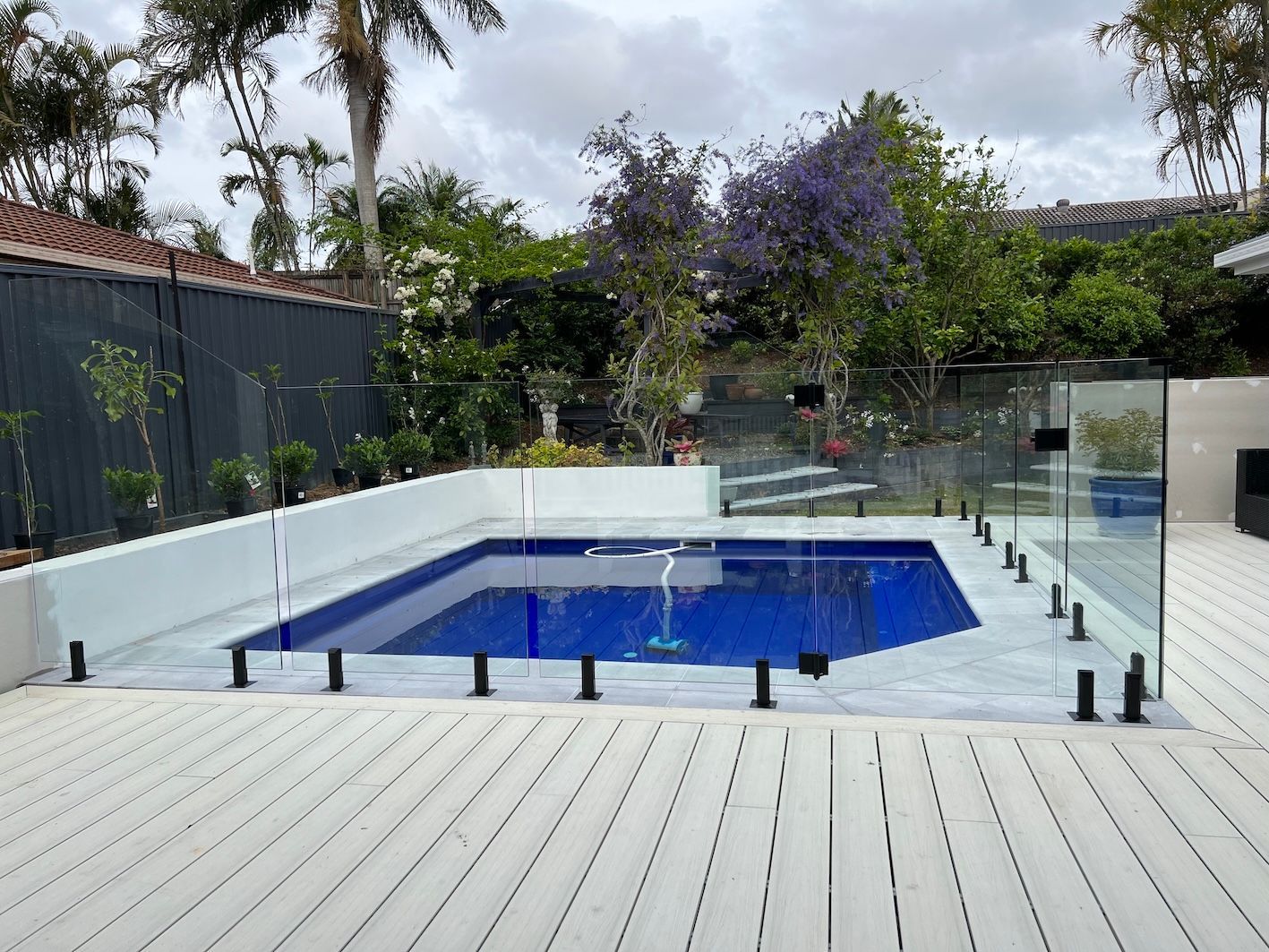 Backyard pool with blue cover, glass fence, and gray deck, surrounded by trees and flowers — Bombs Away Pools in Coolangatta, QLD