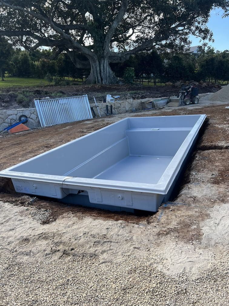 Gray rectangular pool shell set in a dirt excavation, with a large tree in the background.
