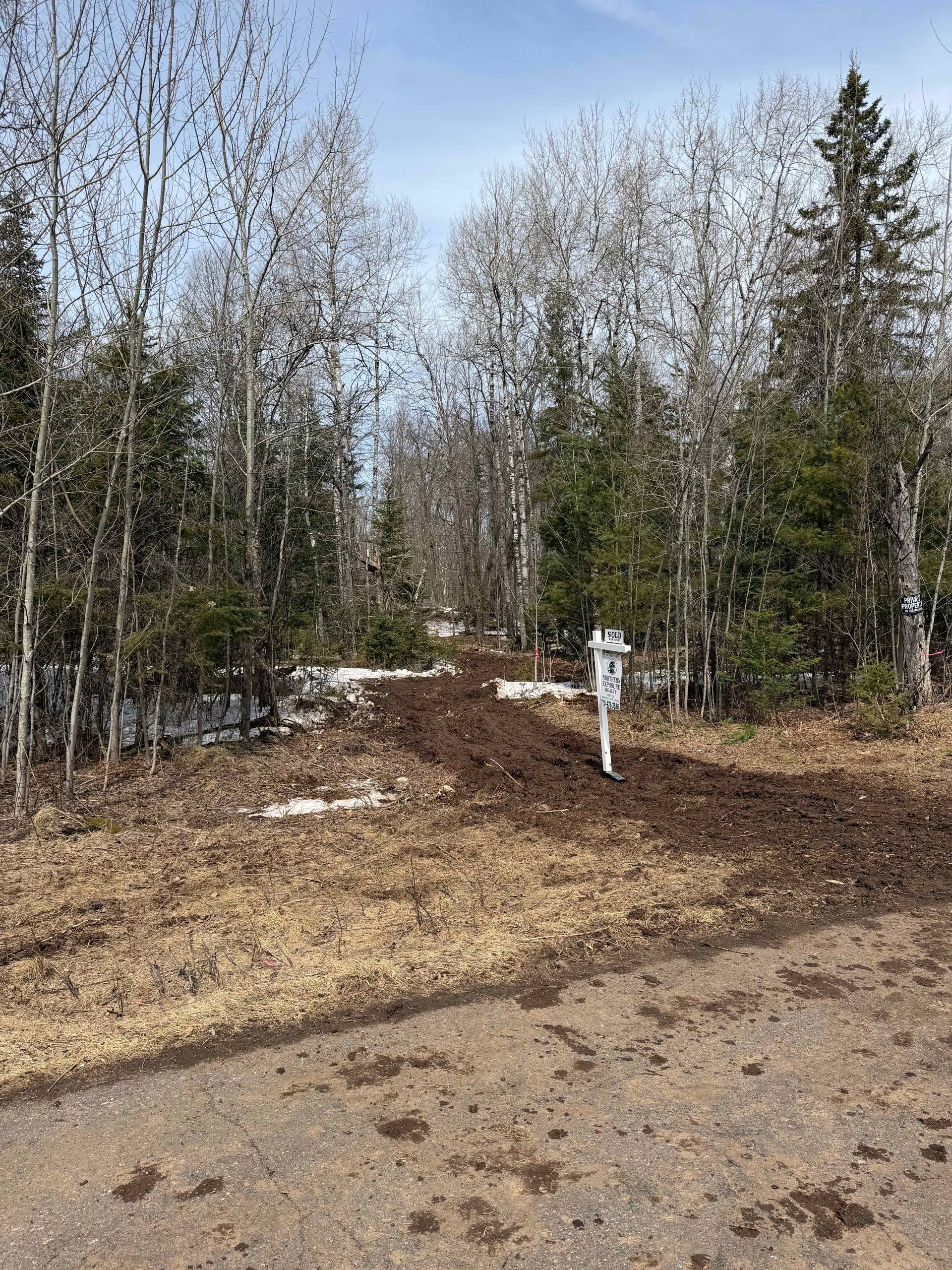 A dirt path leads into a wooded area with patches of snow under tall, leafless trees on a bright, sunny day.