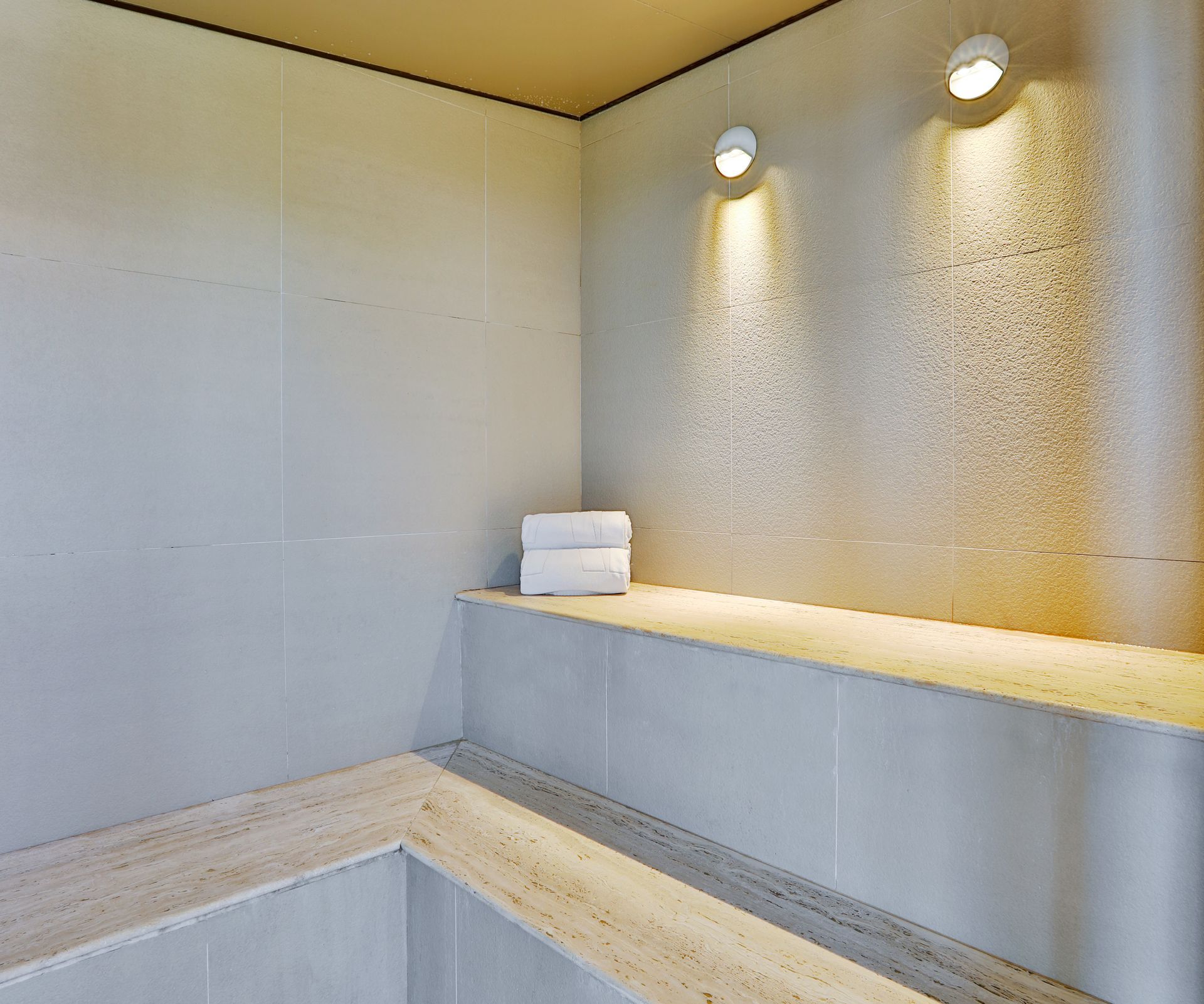 Chuveiro spa moderno com paredes cinza, banco bege e duas luminárias de parede sobre um suporte para toalhas.