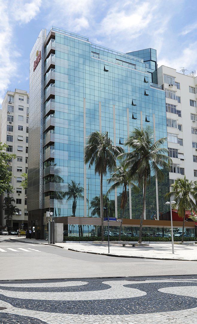 Prédio alto de vidro azul com palmeiras na frente, ao lado de prédios de apartamentos brancos e uma rua com padrões.