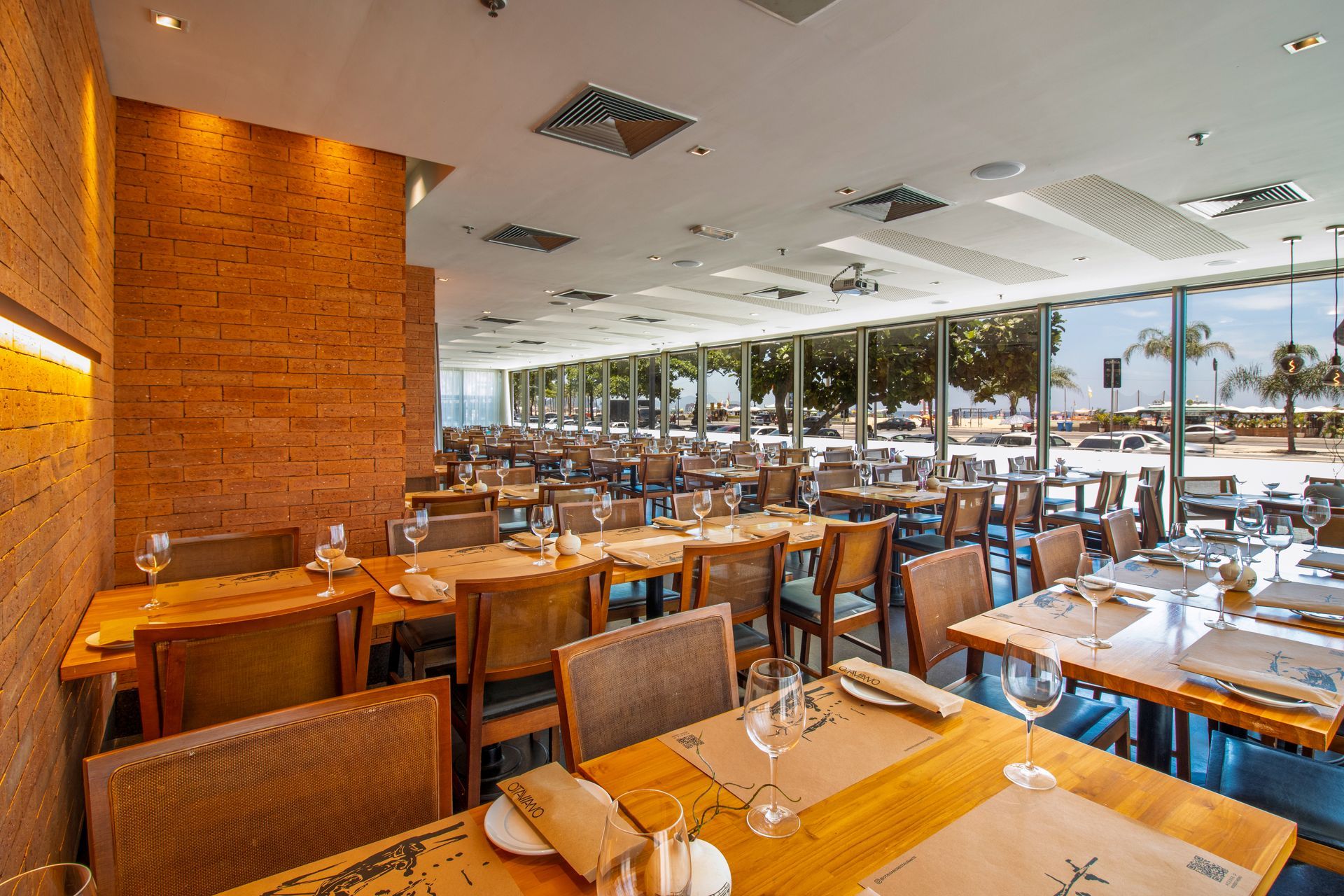 Interior de restaurante moderno com mesas e cadeiras de madeira e grandes janelas com vista para a cidade.