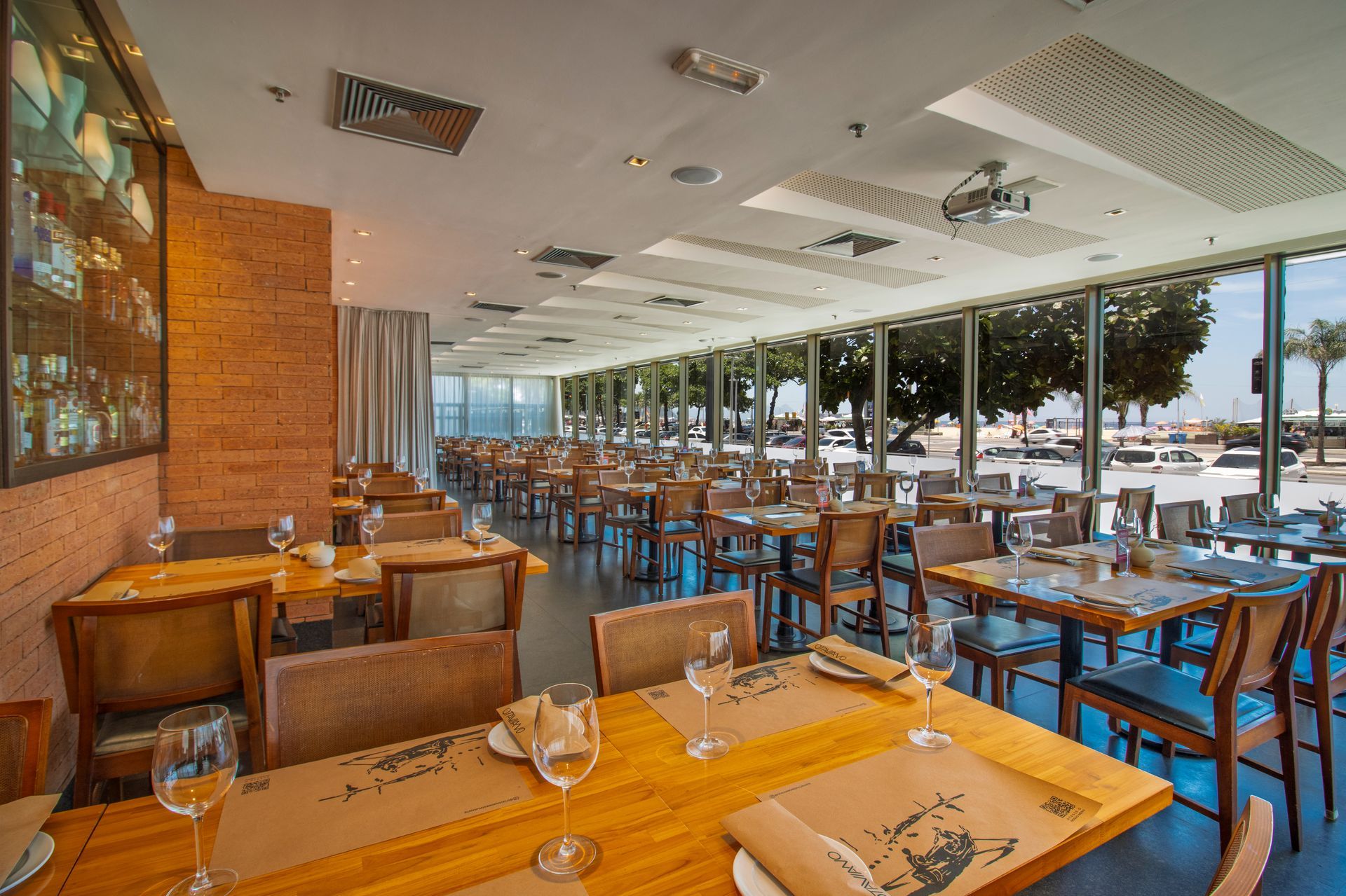 Interior de restaurante moderno com mesas e cadeiras de madeira e grandes janelas com vista para a orla.