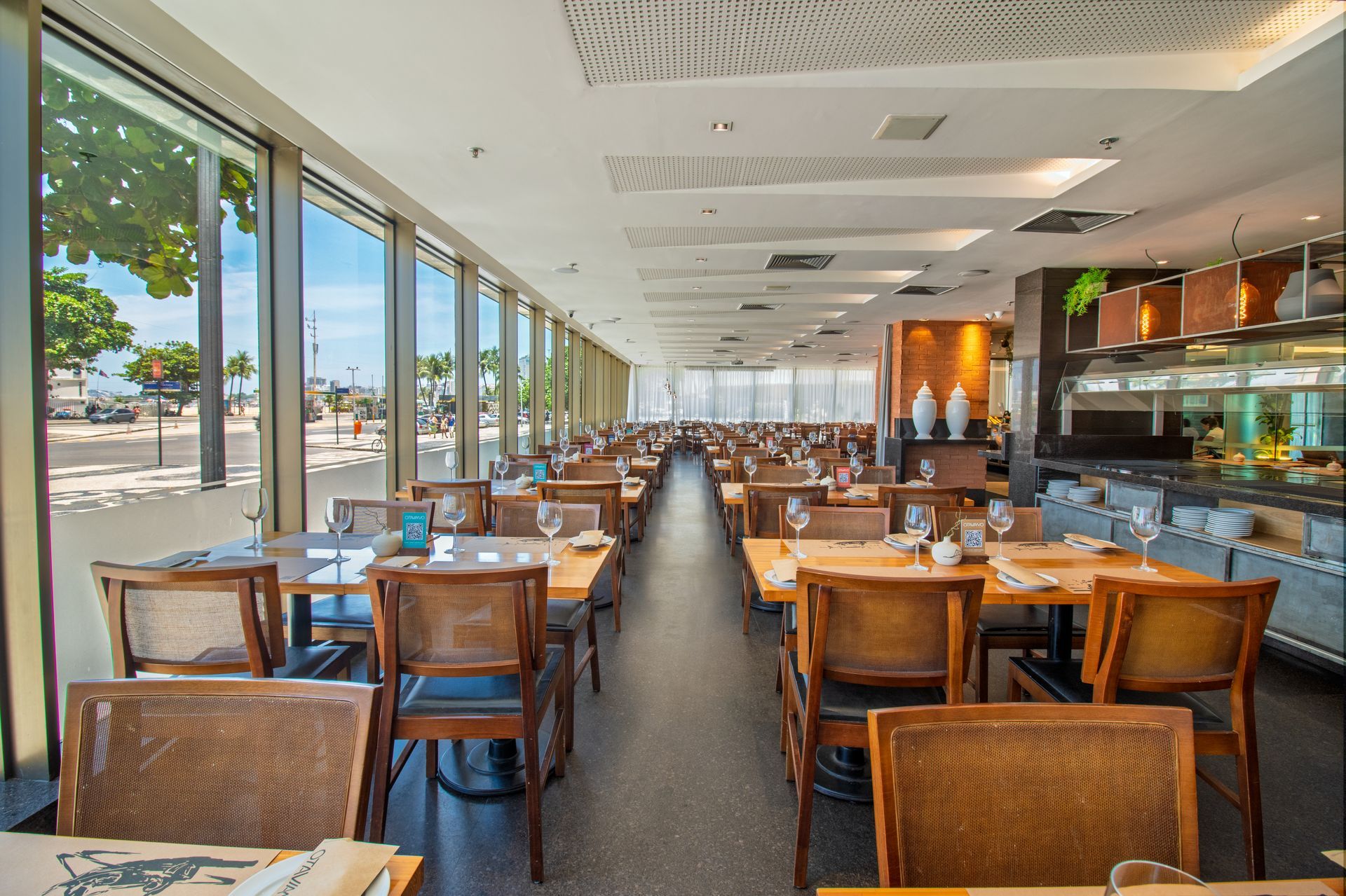 Sala de jantar vazia de um restaurante com mesas de madeira, cadeiras de vime e grandes janelas que deixam entrar a luz do dia.