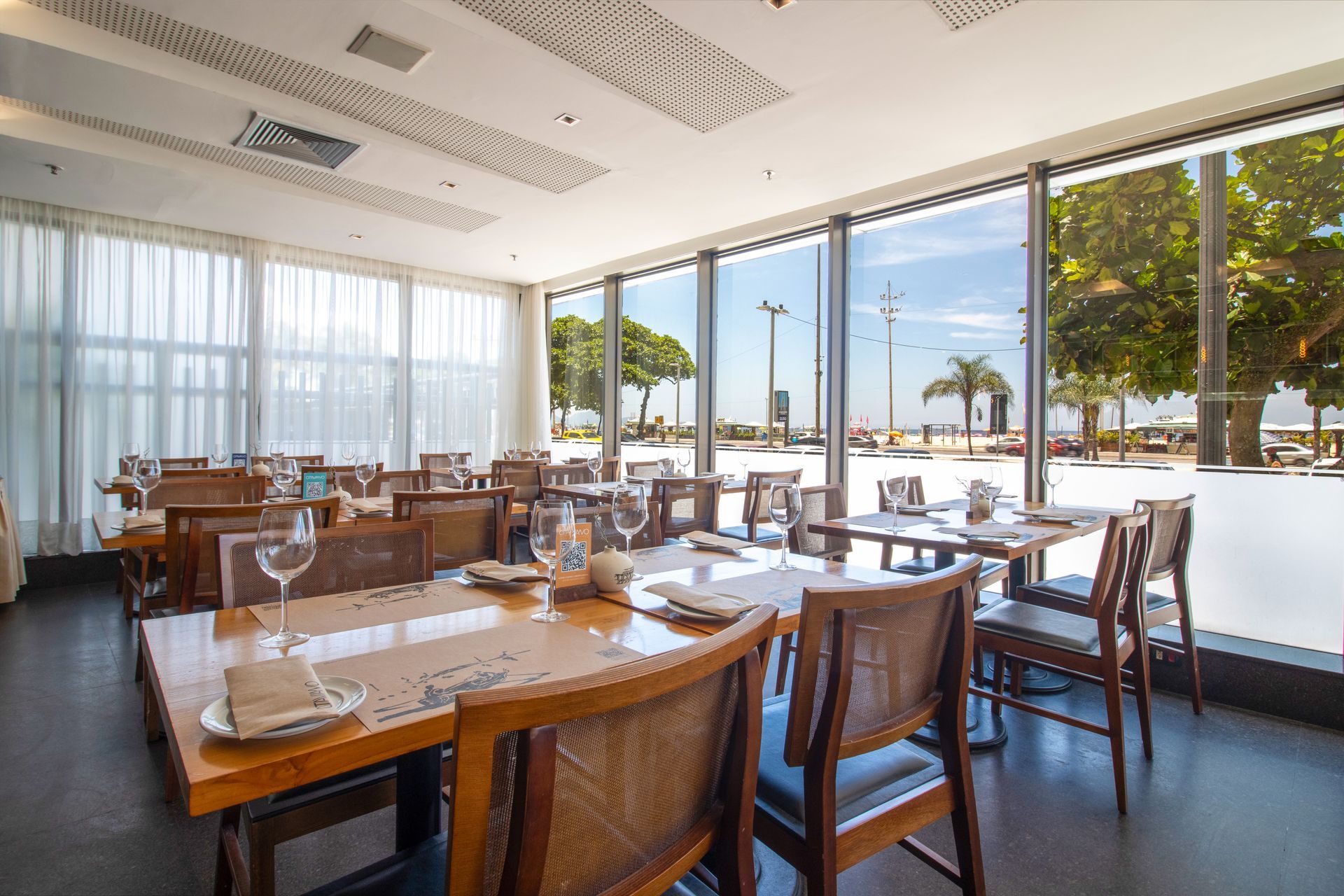 Sala de jantar luminosa do restaurante, com mesas de madeira postas e grandes janelas com vista para uma ensolarada orla marítima.