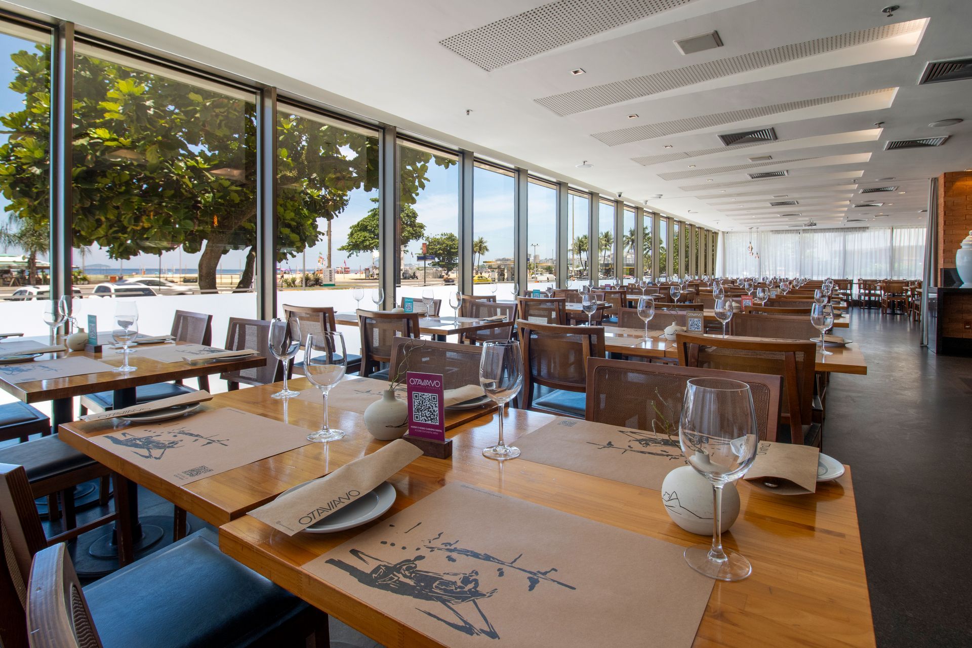 Restaurante luminoso à beira-mar com mesas de madeira, mesas postas e grandes janelas com vista para palmeiras e para o mar.
