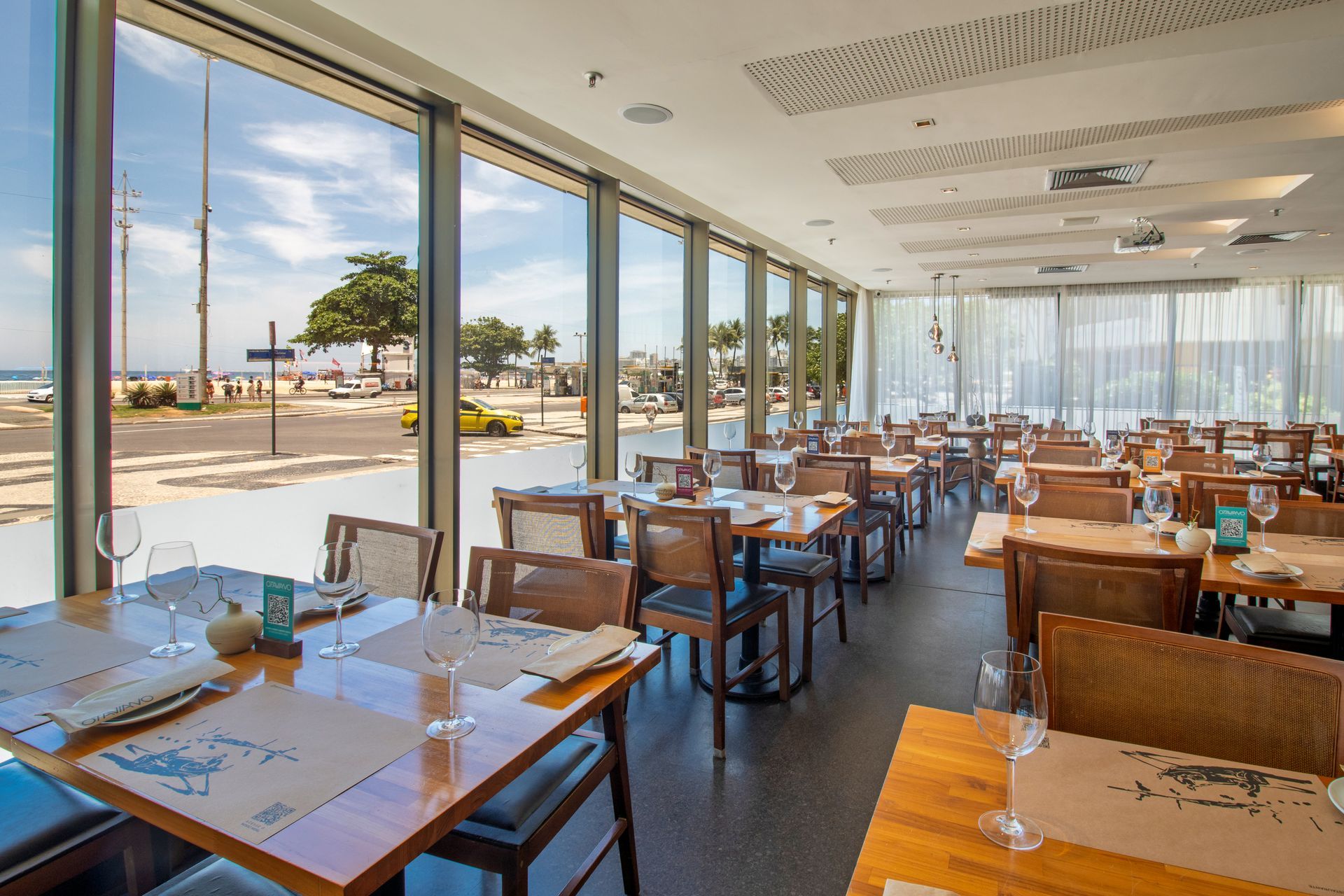 Restaurante luminoso à beira-mar com grandes janelas, mesas de madeira e mesas postas para refeições com vista para o mar.