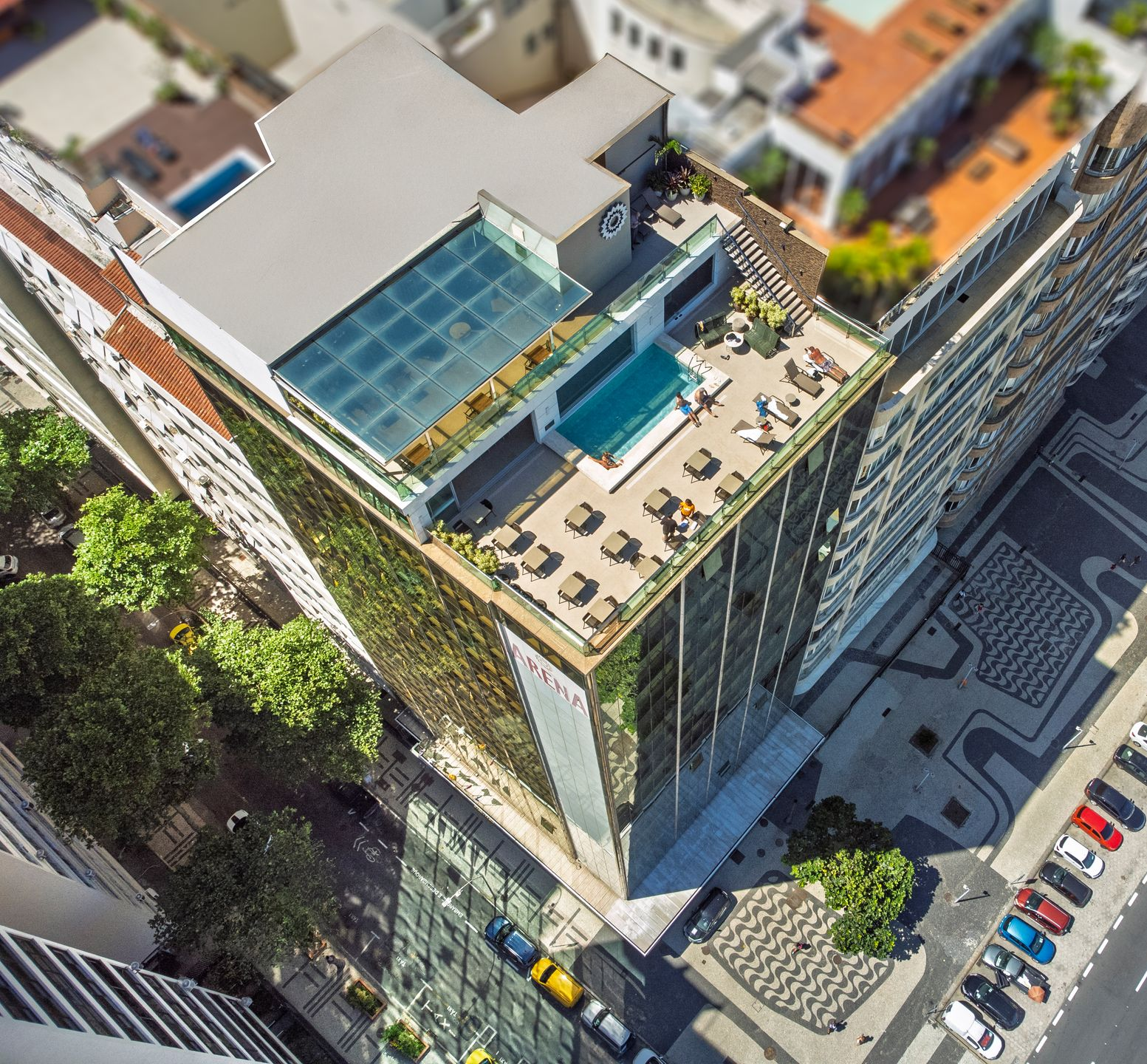 Vista aérea de um terraço alto com piscina e espreguiçadeiras ao lado de prédios e ruas da cidade.