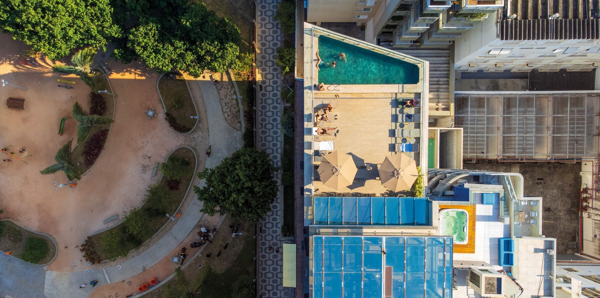 Vista aérea da área da piscina de um resort, ao lado de um caminho circular no jardim e dos edifícios adjacentes.