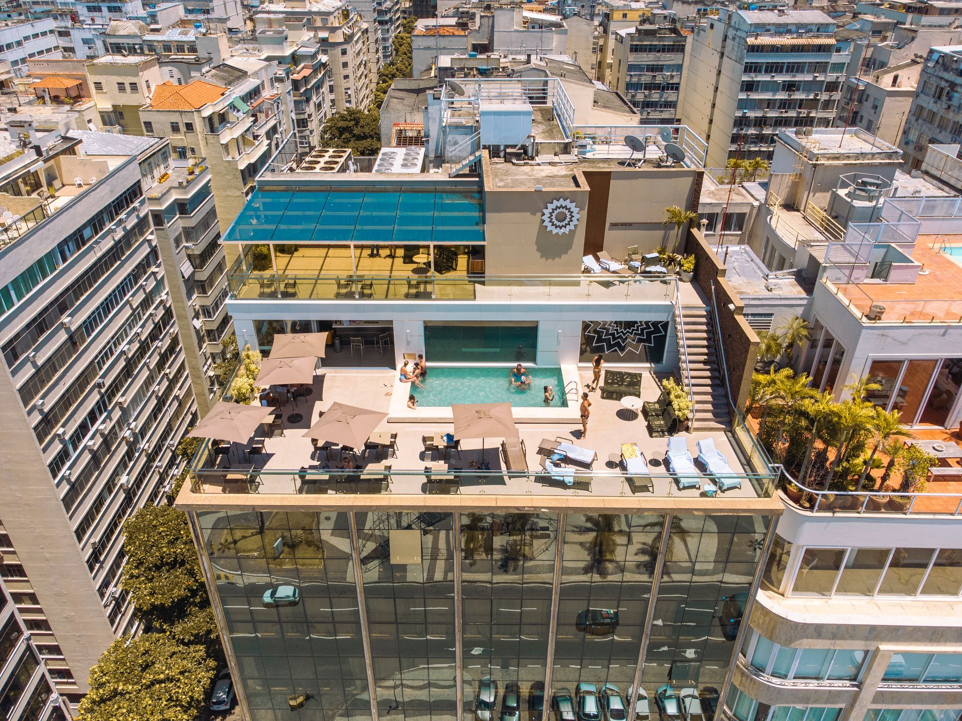 Terraço com piscina na cobertura e espreguiçadeiras no topo de um prédio moderno, cercado por uma densa vegetação urbana.