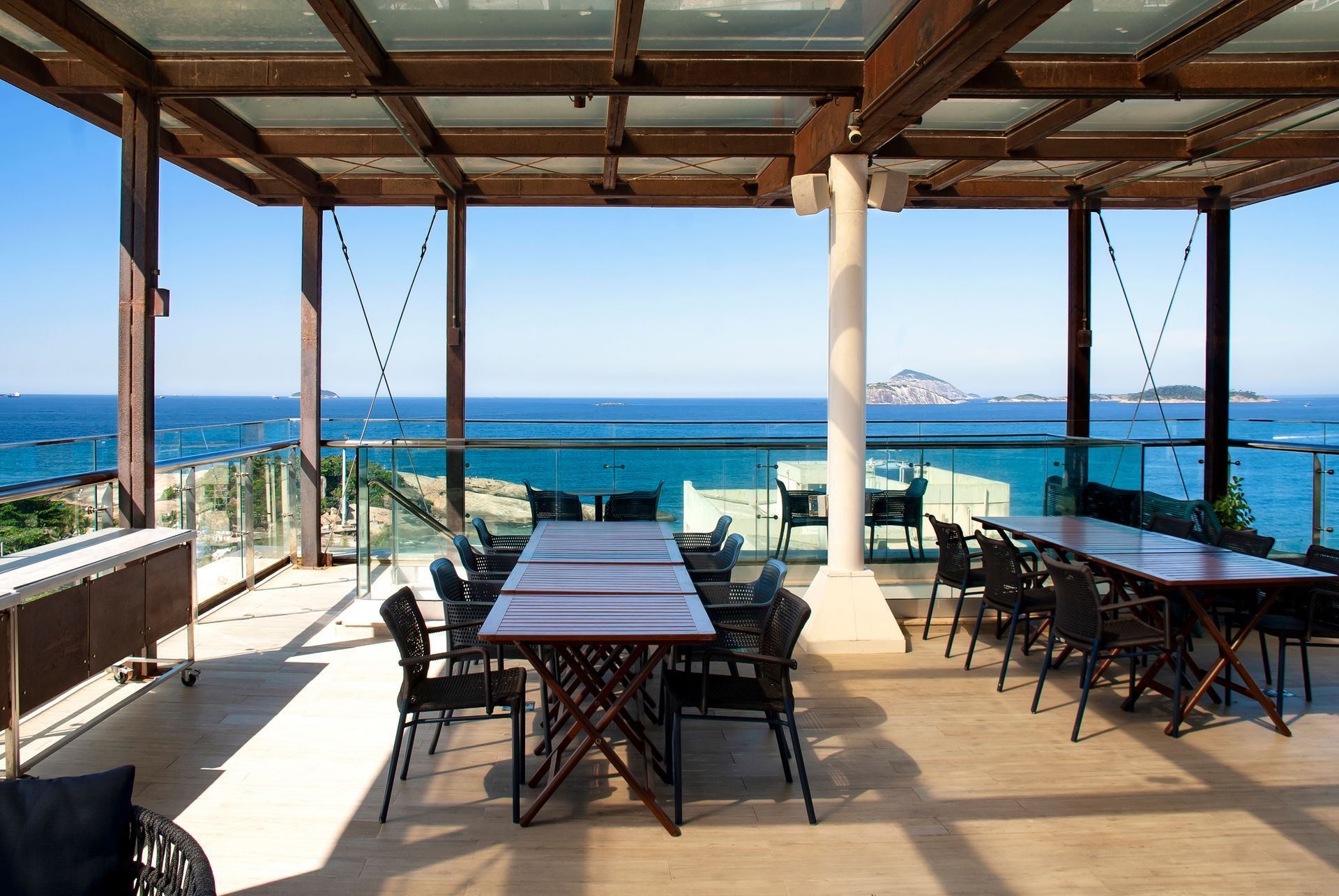 Terraço de restaurante à beira-mar com mesas sob um teto de madeira, com vista para o oceano azul e para a ilha ao longe.