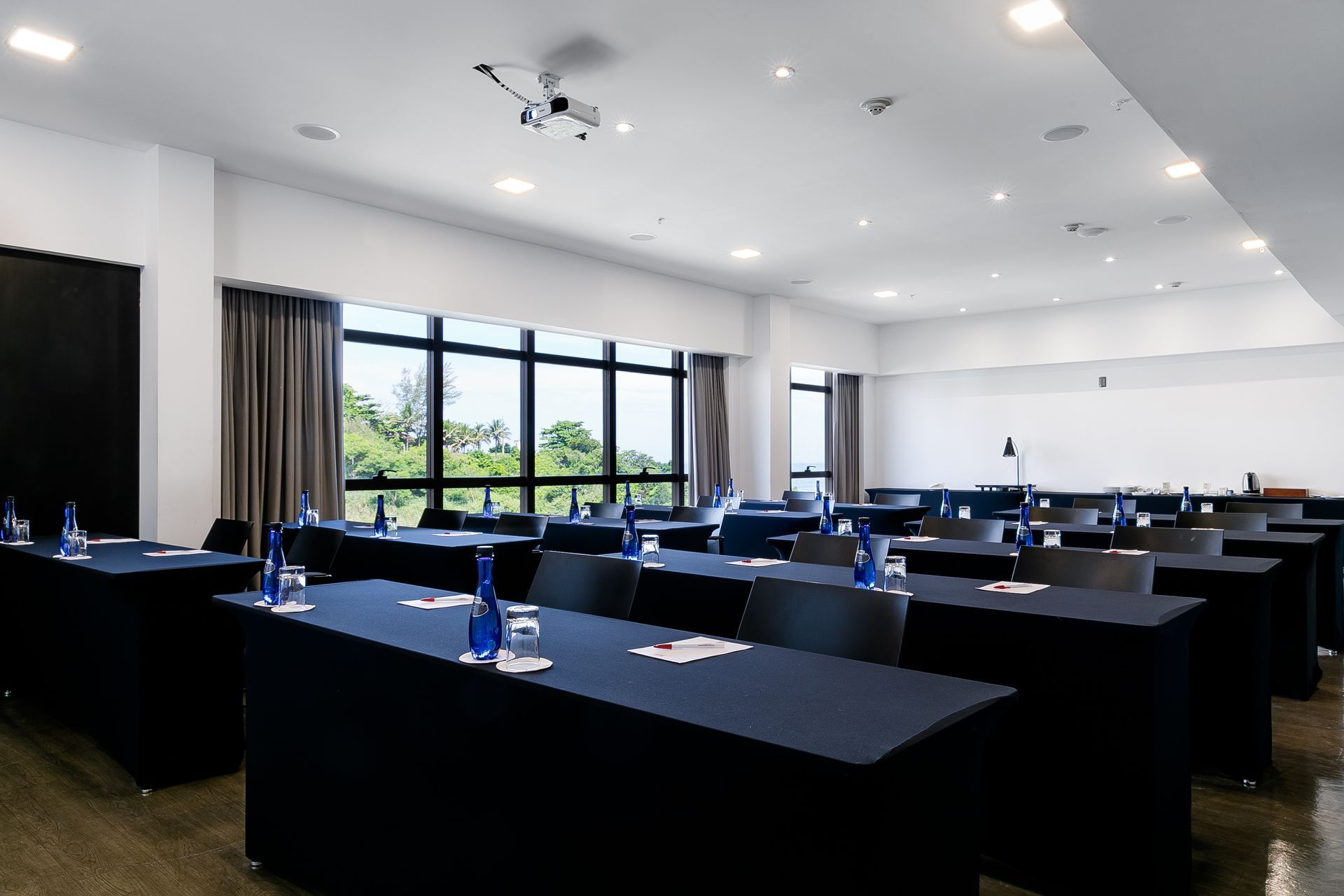 Sala de conferências moderna com mesas pretas, cadeiras azuis e grandes janelas com vista para a vegetação.