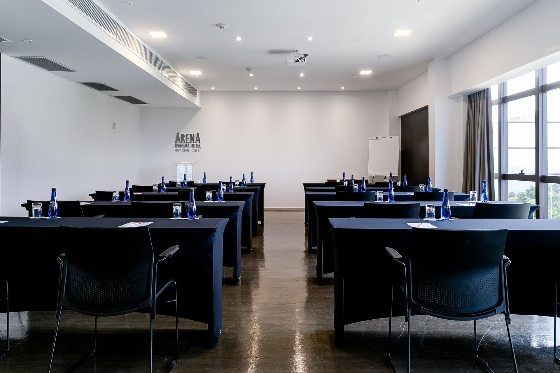 Sala de conferências moderna com mesas e cadeiras pretas dispostas de forma organizada sob iluminação embutida brilhante.